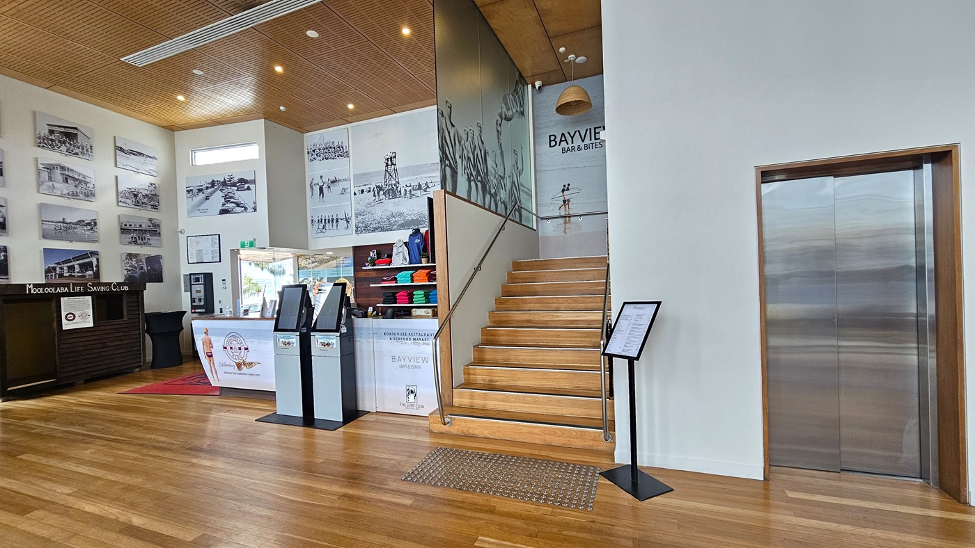 Accessible Lift - Entrance and Reception - The Surf Club Mooloolaba. On the left, there are several black-and-white photographs on the wall above a dark wooden counter with a sign. In the centre, a reception desk with a white banner and two digital kiosks is visible. Behind the desk, shelves display colourful folded clothes. A wooden staircase with a glass railing leads upwards, with a mural on the wall beside it. On the right, a silver lift with a wooden frame is present.