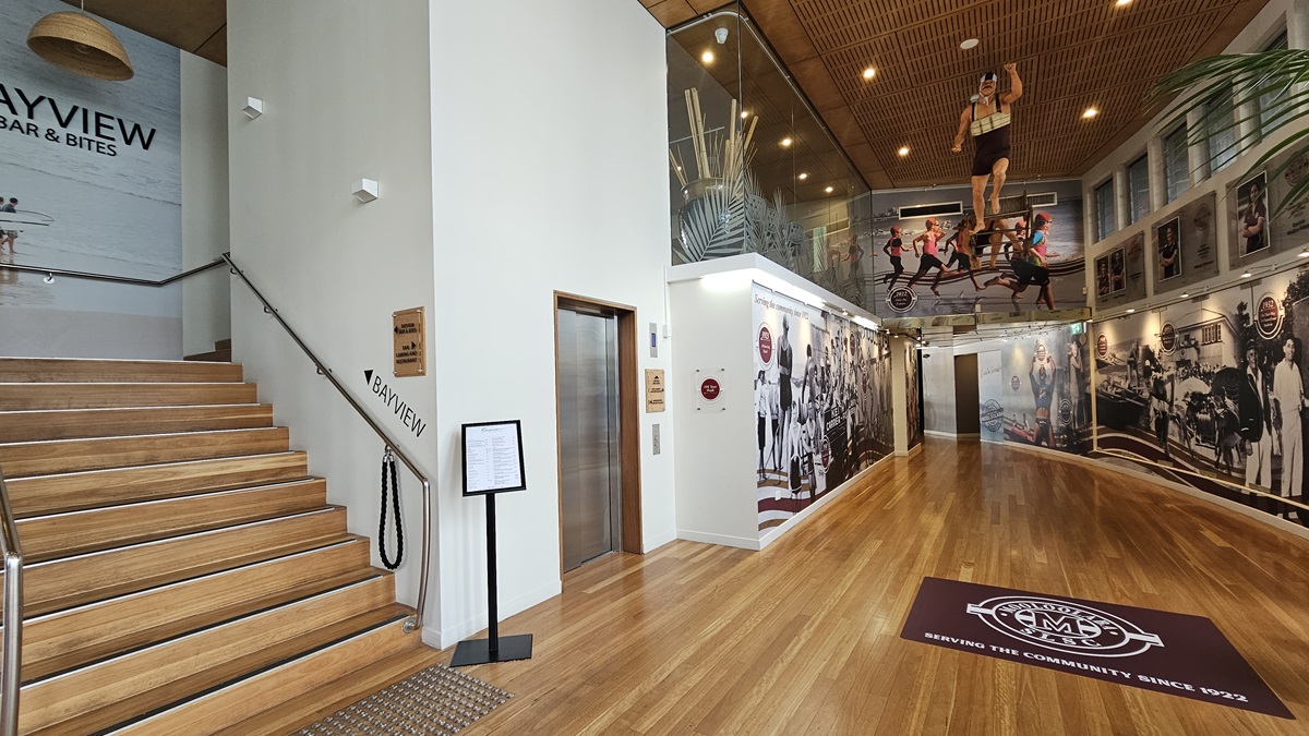 Lift and Corridor - Entrance and Reception - The Surf Club Mooloolaba. A modern interior with wooden flooring and a wooden ceiling featuring recessed lighting. On the left, a staircase with wooden steps leads up to a sign for "Bayview Bar & Bites." An elevator is situated next to the stairs. The right wall is adorned with large, colourful murals depicting various scenes and people. A maroon mat on the floor displays a logo and text. A glass railing above the murals adds a contemporary touch.