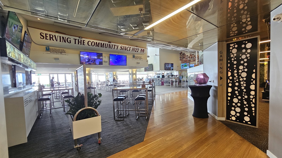 Bar seating area - The Boathouse Restaurant and Bar - The Surf Club Mooloolaba. A spacious interior with a mix of carpeted and wooden flooring. On the left, there are high tables and stools, with a sign above reading "Serving the Community Since 1922". Several screens are mounted on the wall. In the centre, more seating is visible, with a bright, open area in the background. On the right, a decorative panel with circular patterns and a sign indicating "Gaming".