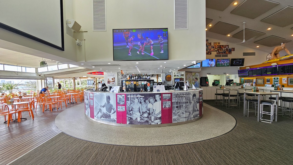 Central Bar - The Boathouse Restaurant and Bar - The Surf Club Mooloolaba. A spacious indoor area with a central circular bar counter featuring black-and-white photos. Above the bar, a large TV screen displays a sports game. To the left, there are orange chairs and tables near large windows, allowing natural light in. On the right, high tables with stools are positioned near a wall with menu boards. The ceiling has recessed lighting and acoustic panels. The floor is a mix of wood and carpeted.