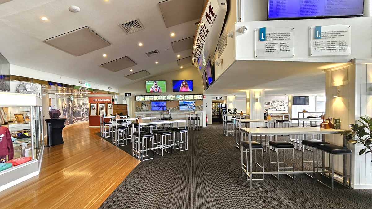 High Table - The Boathouse Restaurant and Bar - The Surf Club Mooloolaba. A spacious interior with a mix of wooden and carpeted flooring. On the left, there are display cases with memorabilia and a curved wall with historical images. In the centre, high tables and stools are arranged, with multiple screens showing sports and other content. The ceiling has recessed lighting and acoustic panels. On the right, more seating is visible, along with informational signs and a plant.
