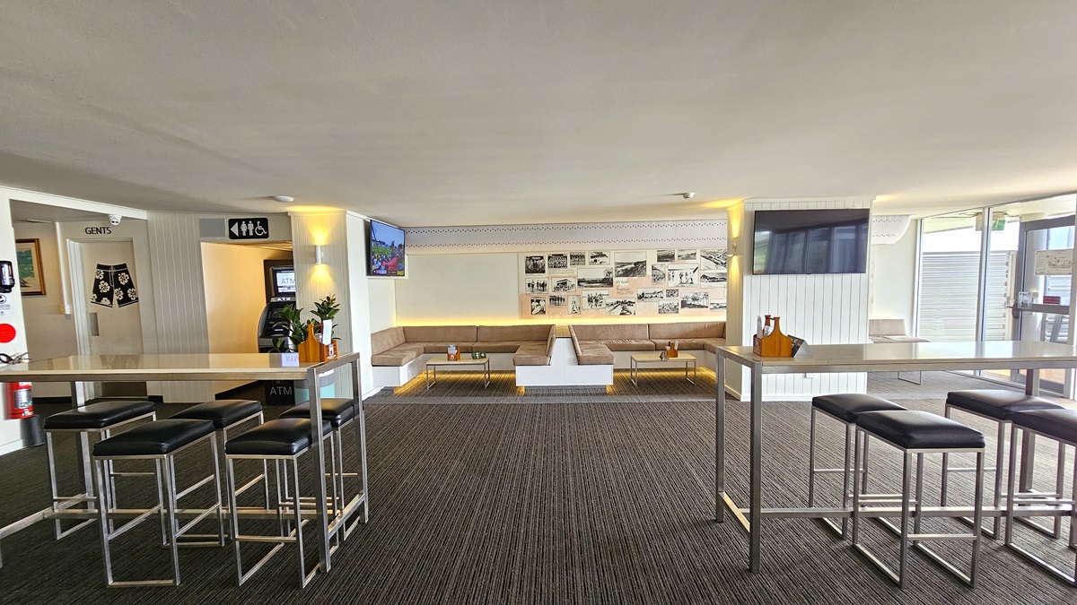 Casual Seating - The Boathouse Restaurant and Bar - The Surf Club Mooloolaba. A modern indoor space with high tables and bar stools on the left and right. The floor is carpeted in a dark pattern. In the centre, there is a seating area with beige cushioned benches arranged in a U-shape, accompanied by small tables. The walls feature framed pictures and a television screen. The ceiling is plain and light-coloured. On the left, there is a doorway marked "Gents" and a sign for restrooms.