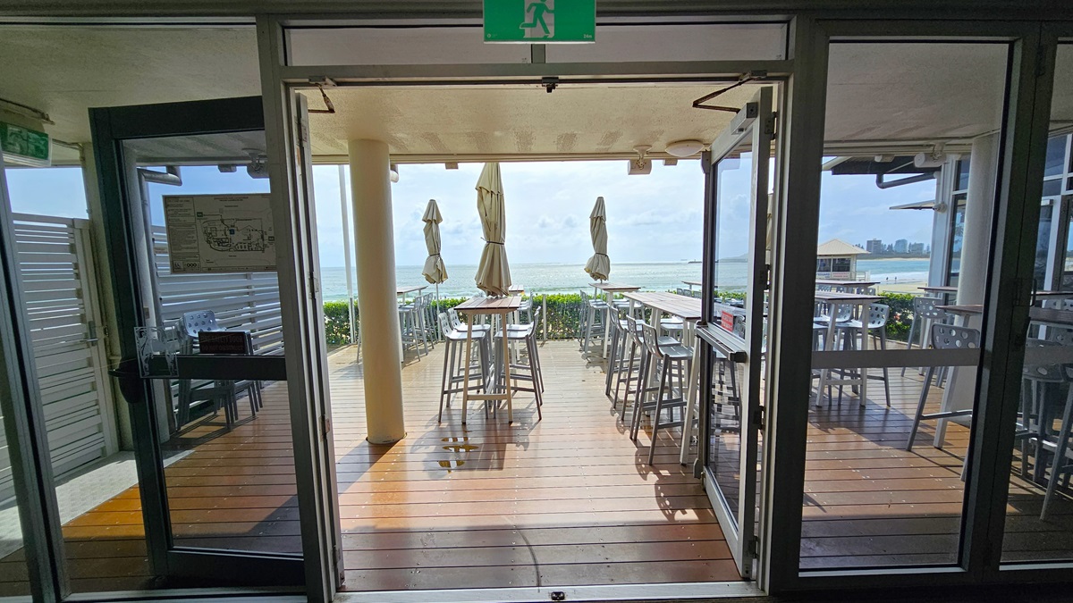 Outdoor sitting area - The Boathouse Restaurant and Bar - The Surf Club Mooloolaba. Glass doors open to a wooden deck with high tables and stools. Closed umbrellas are positioned around the tables. The background features a view of the sea and distant cityscape. The ceiling is light-coloured with a green exit sign above the doors. The floor inside is wooden, matching the deck outside. On the left, there's a wall with a poster and a small table with chairs.