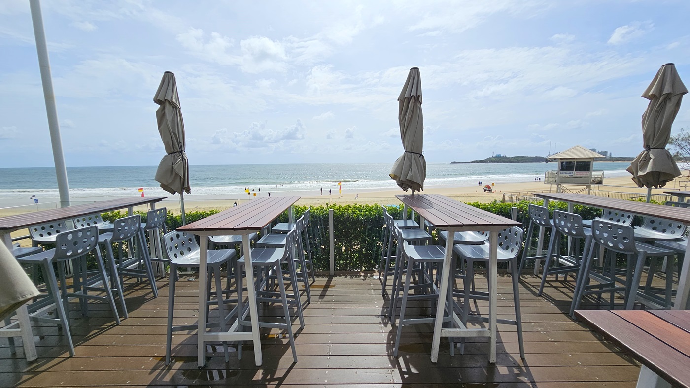 Outdoor deck with views - The Boathouse Restaurant and Bar - The Surf Club Mooloolaba. Wooden deck with high tables and metal stools, some tables have closed umbrellas. The deck overlooks a sandy beach with a few people and a lifeguard tower. The ocean is visible in the background, with gentle waves approaching the shore. A small island or landmass is seen in the distance on the right. The deck is bordered by a low hedge.