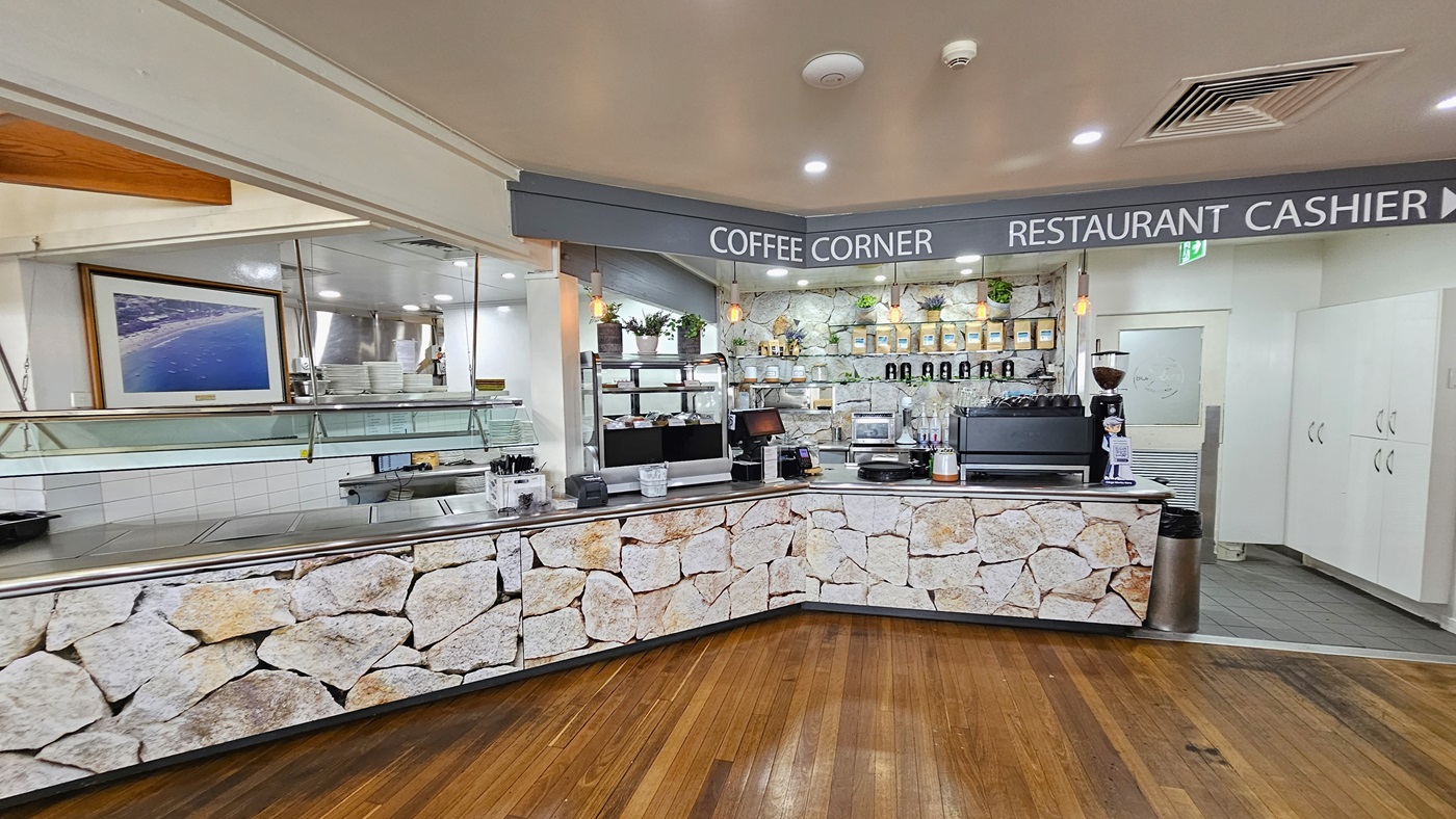 Food order counter - The Boathouse Restaurant and Bar - The Surf Club Mooloolaba. A coffee corner with a stone-patterned counter and wooden flooring. The counter is equipped with coffee machines, a cash register, and various supplies. Shelves behind the counter display coffee bags and potted plants. Overhead, pendant lights illuminate the area. To the left, a framed aerial photograph hangs on the wall. A sign above reads "COFFEE CORNER" and "RESTAURANT CASHIER".