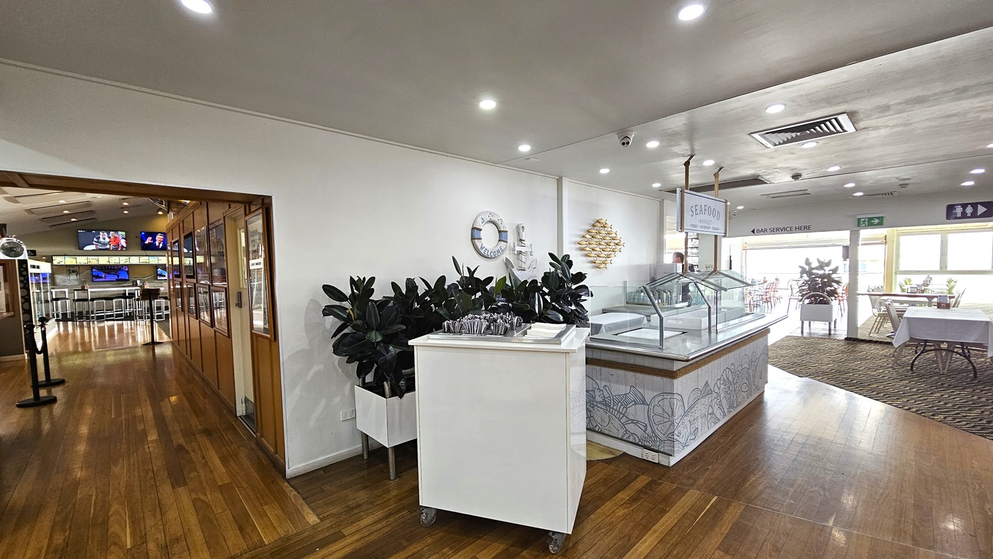 Dining room corridor - The Boathouse Restaurant and Bar - The Surf Club Mooloolaba. A spacious indoor area with wooden flooring. On the left, there's an entrance to another room with multiple screens displaying images. In the centre, a white cart with cutlery is placed next to a large potted plant. On the right, a counter with a glass display and a sign above it. The ceiling is fitted with recessed lights and an air vent. In the background, tables with white tablecloths are near large windows.