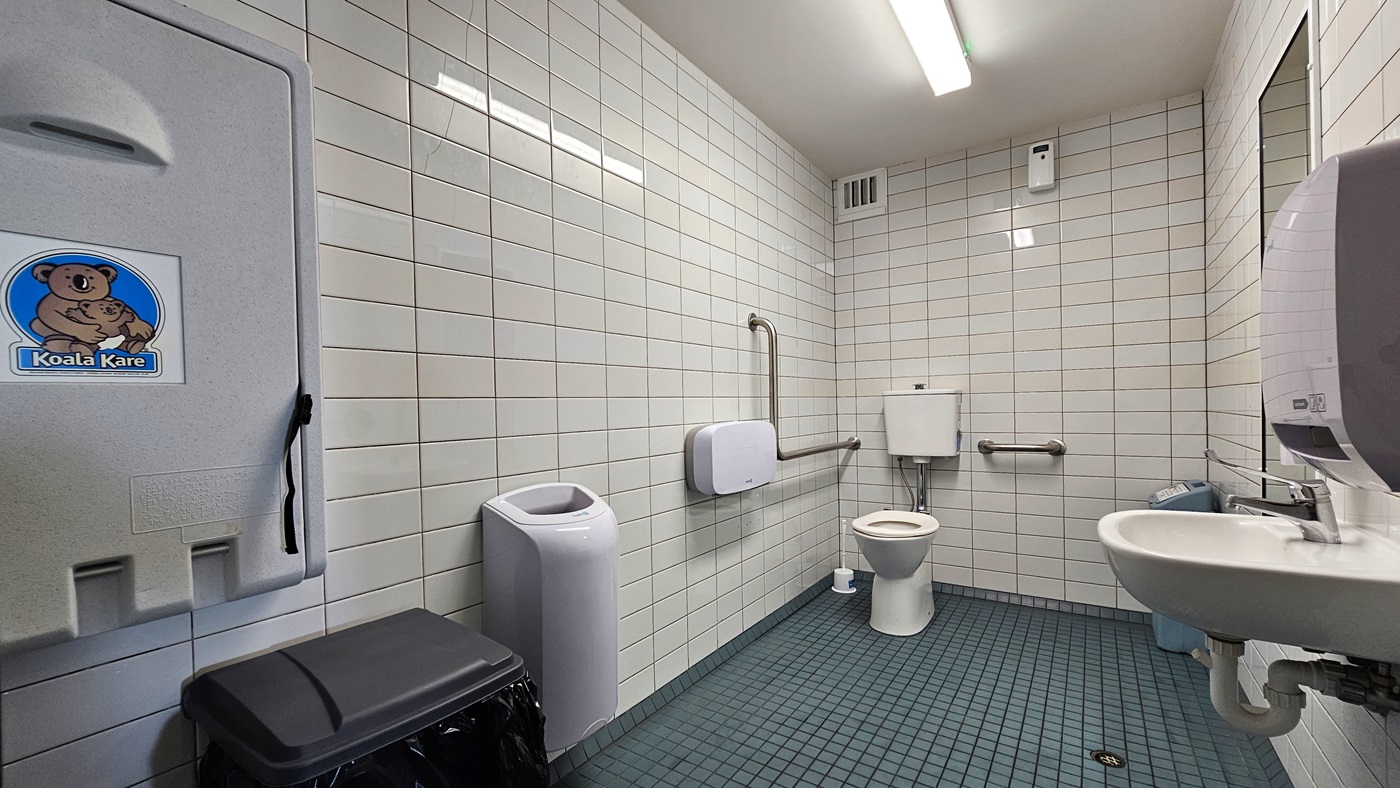 Accessible Bathroom - The Boathouse Restaurant and Bar - The Surf Club Mooloolaba. A public restroom with white tiled walls and a green tiled floor. On the left, a baby changing station with a "Koala Kare" logo is mounted on the wall above a black bin. A white bin is next to it. In the centre, a toilet with a grab bar and a toilet paper dispenser is visible. On the right, a white sink with a soap dispenser is mounted on the wall. A mirror is above the sink. The ceiling has fluorescent lights.
