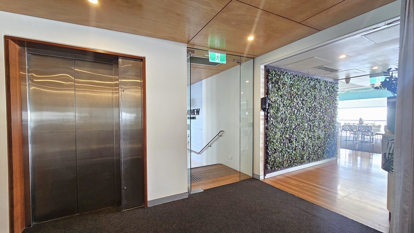Lift and stair access - Bayview Bar and Bites - The Surf Club Mooloolaba. On the left, there is a stainless steel lift with a wooden frame. In the middle, a glass door leads to a stairway with a handrail. On the right, a wall covered in greenery is visible, adjacent to a room with wooden flooring and tables. The ceiling is wooden with recessed lighting, and the floor is carpeted near the lift and transitions to wood near the greenery wall. An exit sign is above the glass door.