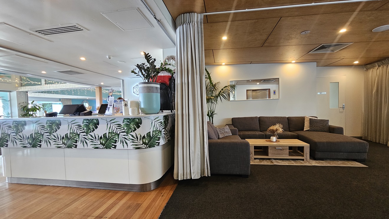 Bar entrance - Bayview Bar and Bites - The Surf Club Mooloolaba. A modern interior with a reception area on the left, featuring a counter decorated with green leaf patterns. Behind the counter are plants and a water dispenser. On the right, a cosy seating area with a large grey sectional sofa and a wooden coffee table adorned with flowers. The floor is a mix of wood and dark carpet, and the ceiling is wooden with recessed lighting. A curtain partially divides the spaces.