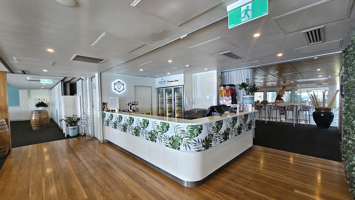 Bar and entry corridor - Bayview Bar and Bites - The Surf Club Mooloolaba. A modern interior with a wooden floor and a white ceiling featuring recessed lighting. In the centre, a counter with a tropical leaf design displays a variety of drinks. Behind the counter, there are glass refrigerators and a neon sign. To the right, high tables and chairs are arranged on a dark carpet. On the left, a wooden barrel and potted plants decorate the space. An emergency exit sign is visible on the ceiling.