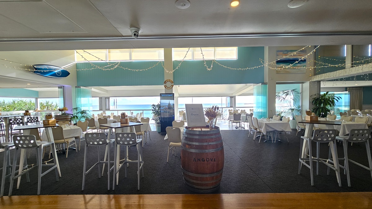 Dining area - Bayview Bar and Bites - The Surf Club Mooloolaba. A spacious indoor dining area with a view of the ocean in the background. The room features high ceilings with string lights and a surfboard decoration. In the centre, there's a wooden barrel with a sign on top, surrounded by white chairs and tables. To the left, there are high tables with stools, and to the right, more dining tables with white tablecloths. Large windows allow natural light to fill the space.