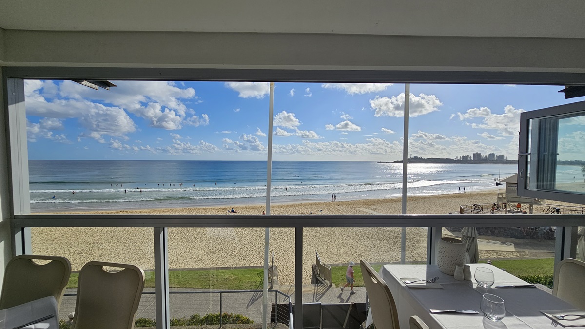 Beach Views - Bayview Bar and Bites - The Surf Club Mooloolaba. A view from a room with large windows overlooking a sandy beach and the ocean. In the foreground, there are dining tables set with white tablecloths, napkins, and glassware. The beach outside is populated with people enjoying the water and sand. In the distance, a city skyline is visible on the right. The ceiling is plain, and the floor is not visible.