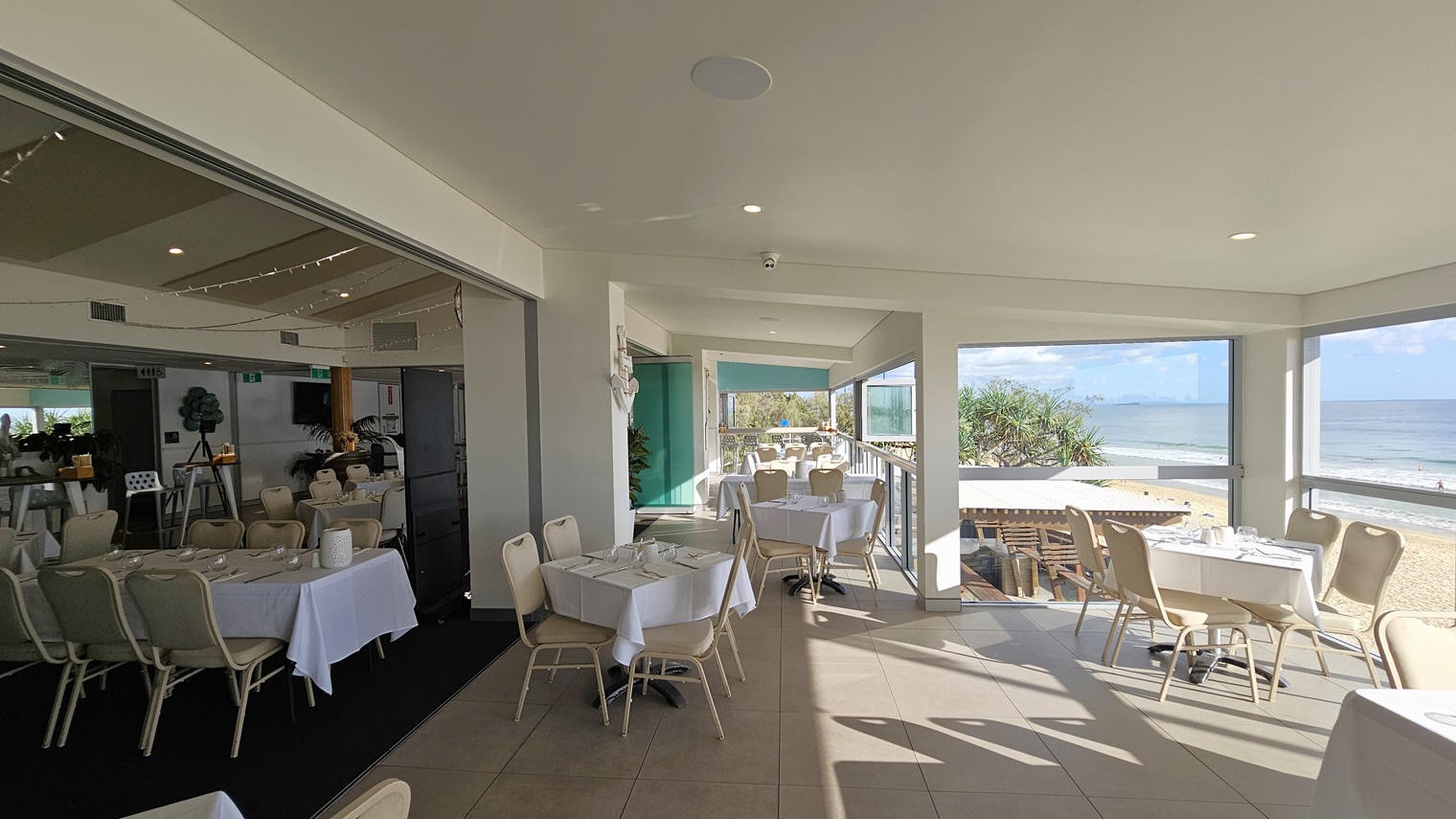 Dining with views - Bayview Bar and Bites - The Surf Club Mooloolaba. A bright, open dining area with tables covered in white tablecloths and beige chairs. On the left, there's an indoor section with more tables and a dark floor. The right side features large windows offering a view of the beach and ocean. The ceiling is light-coloured with recessed lighting, and the floor is tiled. The outdoor area has additional seating with a view of the sea and greenery.