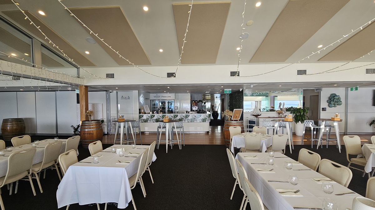 Formal dining - Bayview Bar and Bites - The Surf Club Mooloolaba. A spacious dining area with tables covered in white tablecloths and set with cutlery and glasses. Beige chairs surround the tables. In the background, a bar area with a white counter and decorative items is visible. Barrels and high tables with stools are placed around the room. The ceiling features recessed lighting and string lights, while the floor is a mix of black carpet and wooden panels.