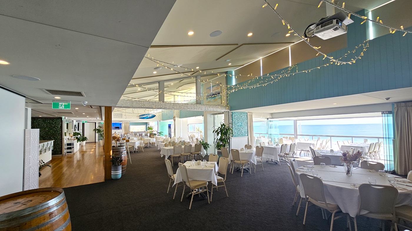 Indoor Dining - Bayview Bar and Bites - The Surf Club Mooloolaba. A spacious indoor venue with a dark carpeted floor and a light-coloured ceiling adorned with string lights. On the left, there are wooden barrels and a wooden floor leading to a bar area with potted plants. In the centre, round tables covered with white tablecloths and beige chairs are arranged neatly. The right side features large windows with views of the ocean, allowing natural light to fill the room.
