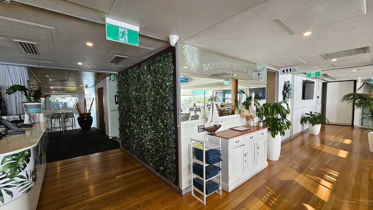 Amenities Corridor Access - Bayview Bar and Bites - The Surf Club Mooloolaba. A modern interior with wooden flooring and a ceiling featuring recessed lighting. On the left, there's a curved counter with a tropical leaf design, leading to a dark carpeted area with large vases and plants. The centre has a wall with greenery and a white cabinet topped with decorative items. On the right, a corridor with potted plants and a wall-mounted TV. An exit sign is visible above.