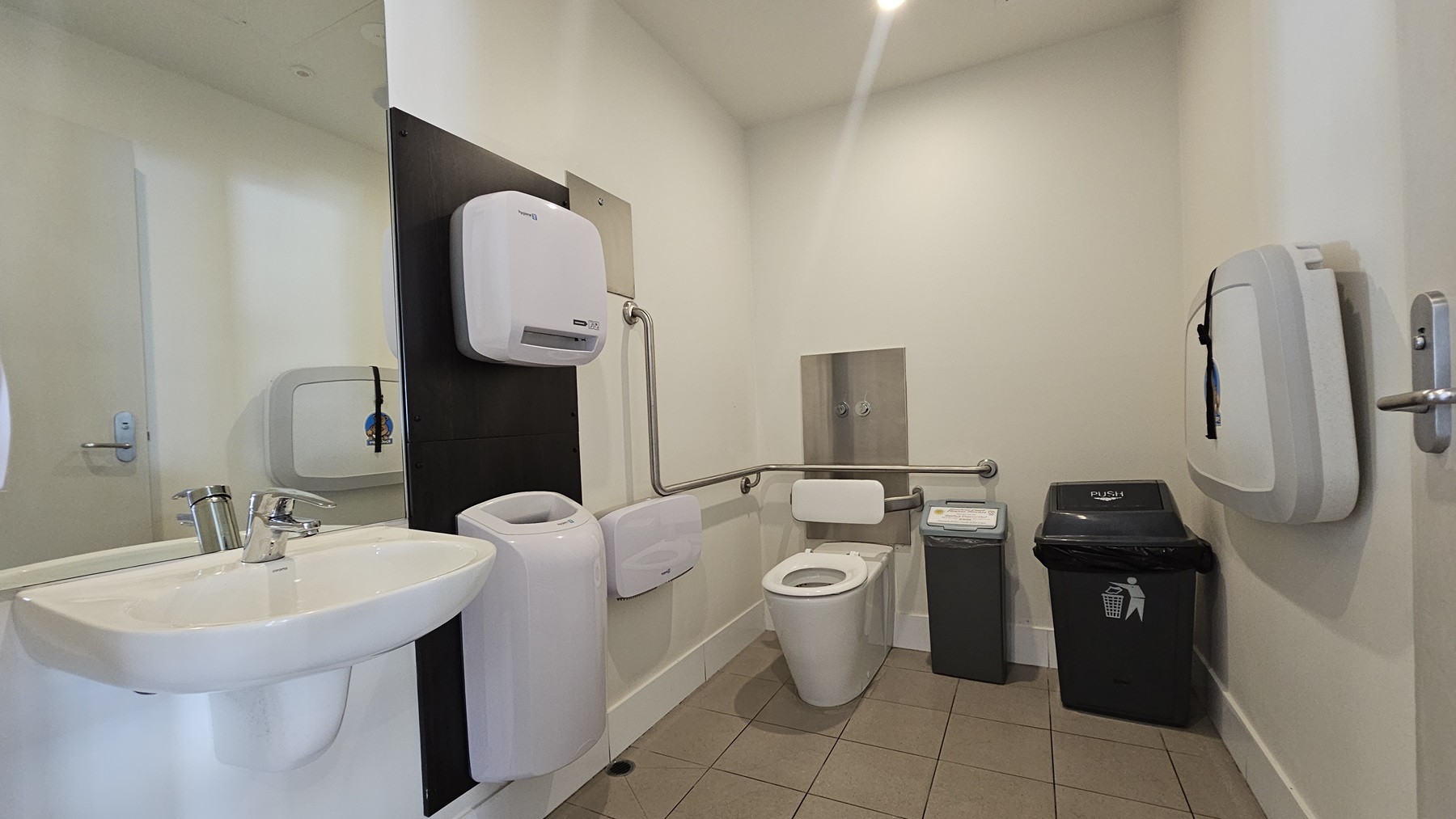 Accessible Bathroom - Bayview Bar and Bites - The Surf Club Mooloolaba. A public restroom with a white sink and chrome tap on the left. Above the sink is a mirror, and next to it, a hand dryer is mounted on a black panel. The toilet is in the centre, equipped with a support rail. On the right, there is a nappy changing station and two bins, one for general waste and another for sanitary disposal. The floor is tiled in grey, and the walls are painted white.