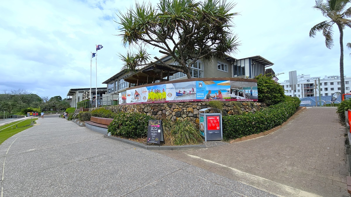 Step free walkway - Mooloolaba Beach Access - The Surf Club Mooloolaba. A paved walkway curves to the left, bordered by grass and a low stone wall. On the right, a building with a modern design is partially obscured by trees and bushes. Colourful banners are displayed along the wall, and a signboard stands near the path. A palm tree is visible on the right, and in the background, there are more buildings. The sky is overcast.