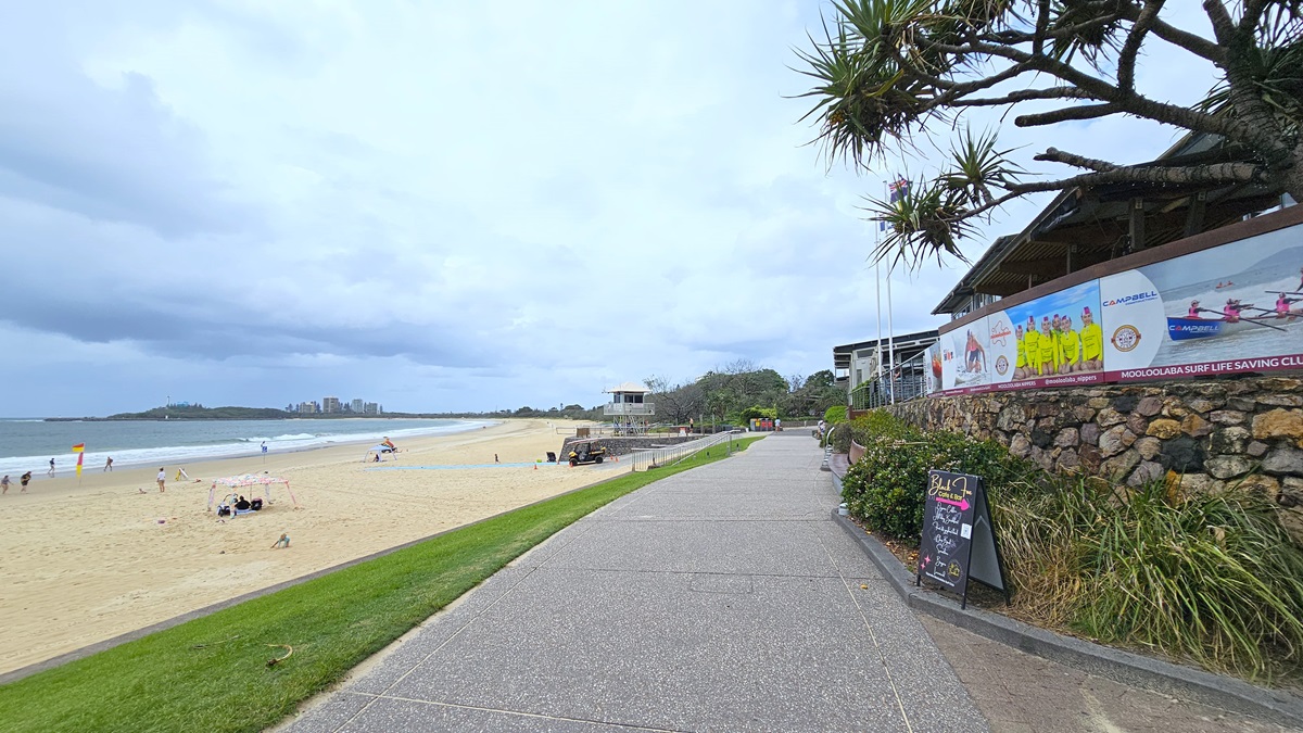 Flat concrete path - Mooloolaba Beach Access - The Surf Club Mooloolaba. A beach scene with a sandy shore on the left, where people are scattered along the water's edge. The ocean stretches into the distance with a city skyline visible on the horizon. On the right, a paved walkway runs parallel to the beach, bordered by a stone wall and greenery. A building with a large sign and a chalkboard stands beside the path. Overhead, tree branches extend into the frame.