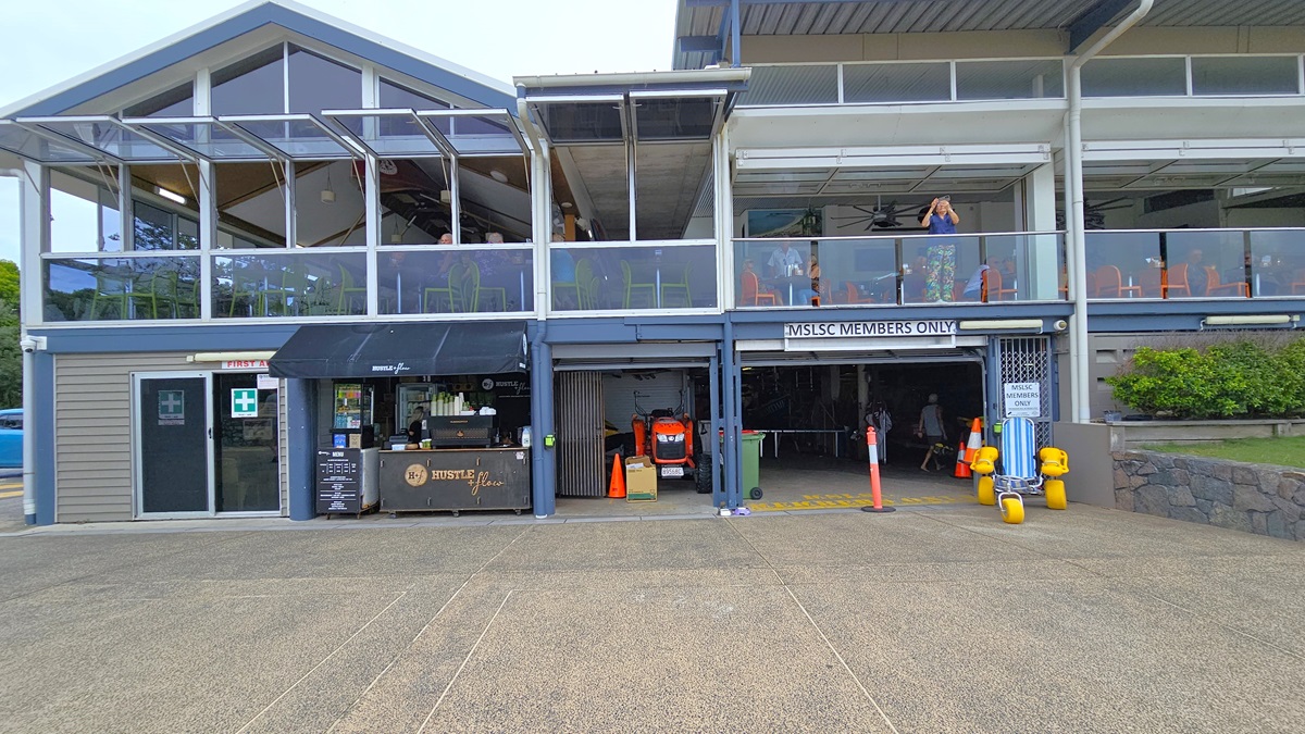 Accessible Beach Wheelchair Collection Point Wide Angle - Mooloolaba Beach Access - The Surf Club Mooloolaba. Two-storey building with a modern design. The ground floor has a small café with a black awning and a menu board. Next to it is a garage area marked "MSLSC Members Only," with orange cones and a small vehicle inside. The upper floor features a glass-enclosed balcony with tables and chairs. The building has a sloped roof with large windows. The foreground has a textured pavement.