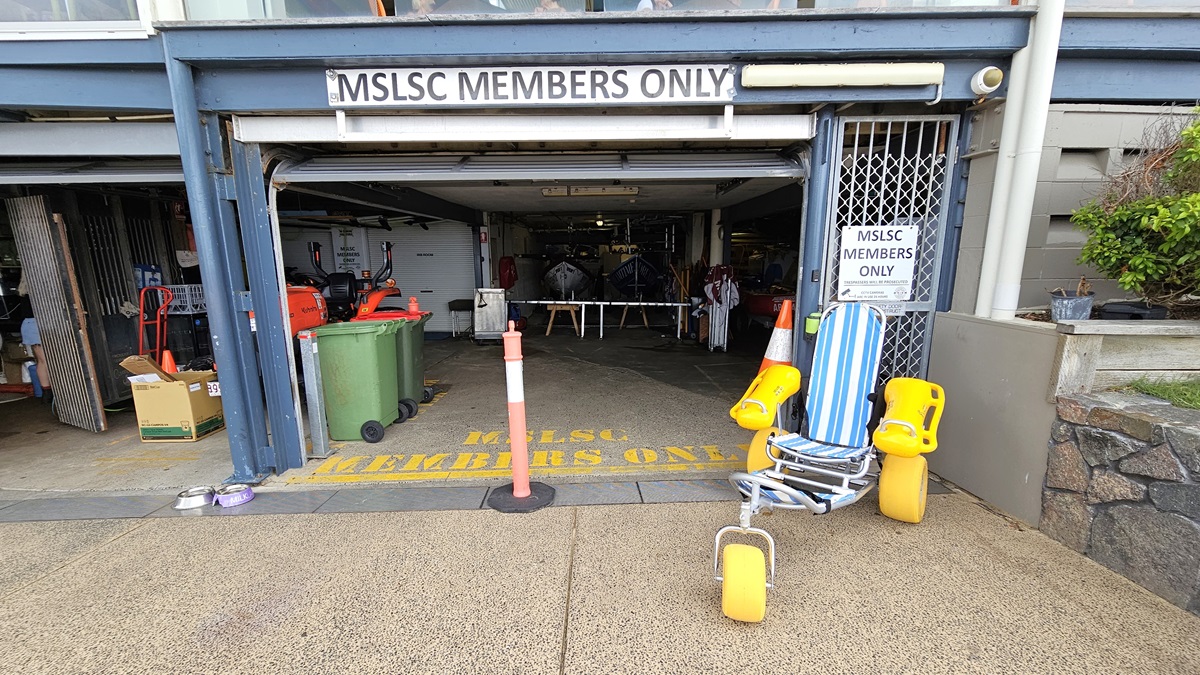Accessible Beach Wheelchair Collection Point - Mooloolaba Beach Access - The Surf Club Mooloolaba. Two-storey building with a modern design. The ground floor has a small café with a black awning and a menu board. Next to it is a garage area marked "MSLSC Members Only," with orange cones and a small vehicle inside. The upper floor features a glass-enclosed balcony with tables and chairs. The building has a sloped roof with large windows. The foreground has a textured pavement.