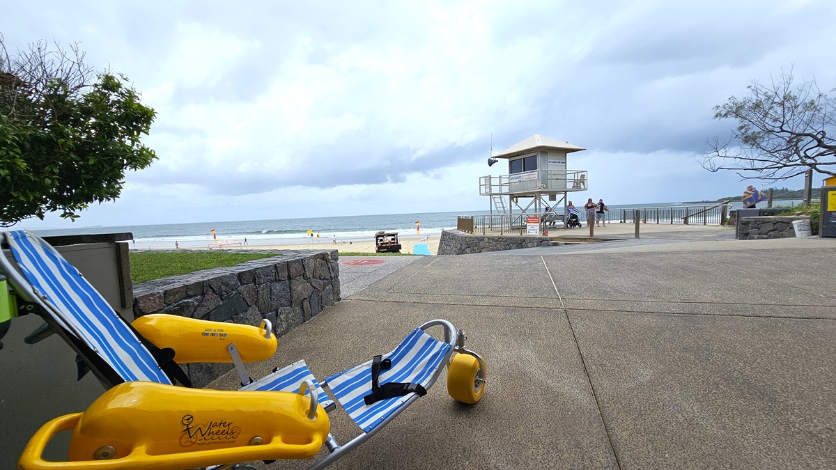 Accessible Beach Wheelchair - Mooloolaba Beach Access - The Surf Club Mooloolaba. A beach scene with a striped blue and white beach wheelchair with yellow wheels on the left. In the middle, a stone wall separates the walkway from the sandy beach. A lifeguard tower is visible in the background, with a few people nearby. The ocean is in the distance, and the sky is overcast. On the right, a tree with sparse branches and a small yellow structure are visible. The ground is step free concrete.