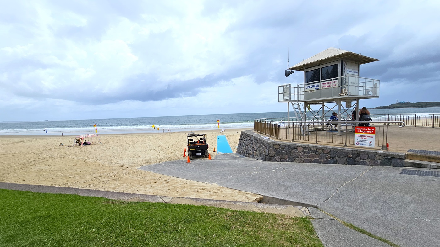 Accessible Beach Matting - Mooloolaba Beach Access - The Surf Club Mooloolaba. A sandy beach with a lifeguard tower on the right, elevated on metal stilts with a railing around it.  A small vehicle is parked on a concrete path leading to the beach, marked by orange cones. A blue accessible beach mat extends from the path onto the sand. In the background, the ocean is visible with gentle waves. A grassy area is in the foreground on the left. Signs are posted near the lifeguard tower.