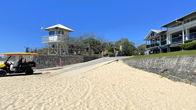 Ramp Access - Mooloolaba Beach Access - The Surf Club Mooloolaba. A sandy beach in the foreground leads up to a concrete ramp. On the left, a lifeguard tower with a sloped roof is elevated on a platform. A small vehicle with a yellow canopy is parked nearby. On the right, there are modern buildings with balconies overlooking the beach. Trees and greenery are visible in the background under a clear blue sky.