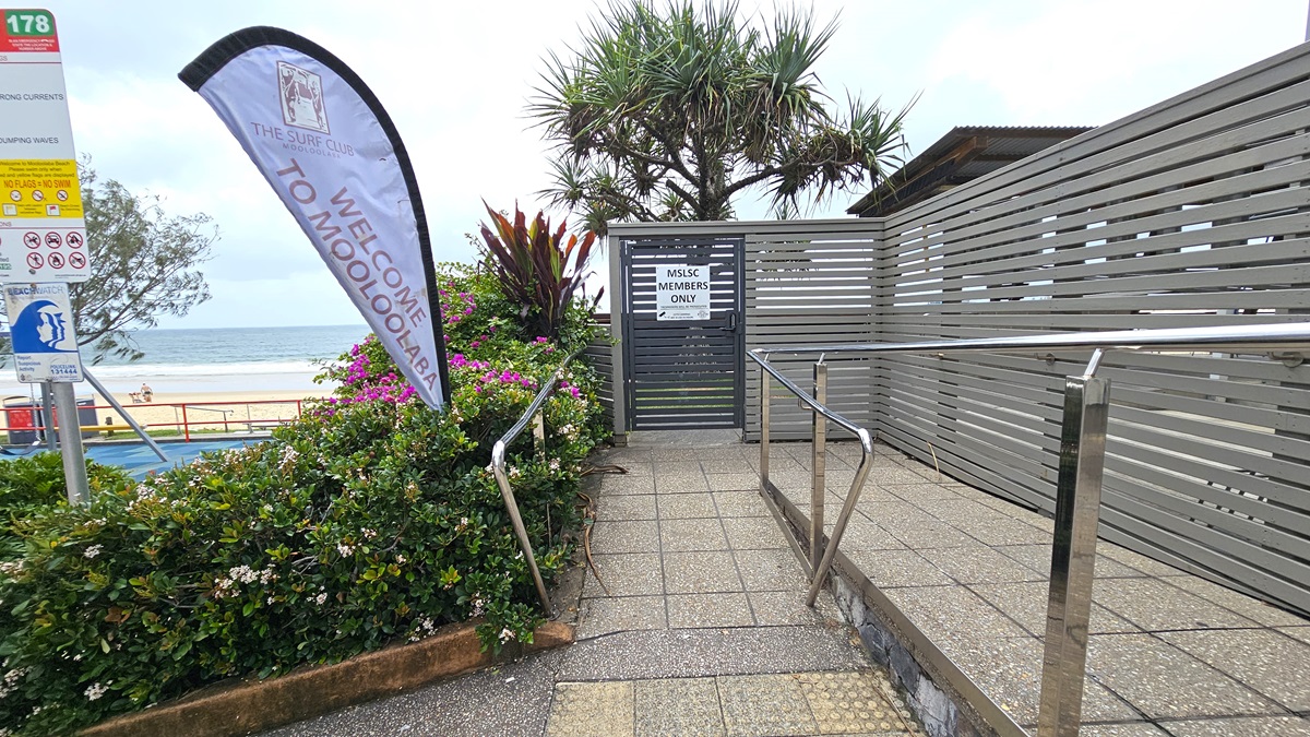 Ramp Access - ANZAC Dawn Service - The Surf Club Mooloolaba. A pathway with a metal railing leads to a gate marked "MSLSC Members Only". On the left, a flag reads "Welcome to Mooloolaba" next to a sign with various symbols. Lush greenery with pink and white flowers lines the path. In the background, the ocean and beach are visible, with a tree providing some shade. The ground is paved with textured tiles, and a wooden fence runs along the right side.