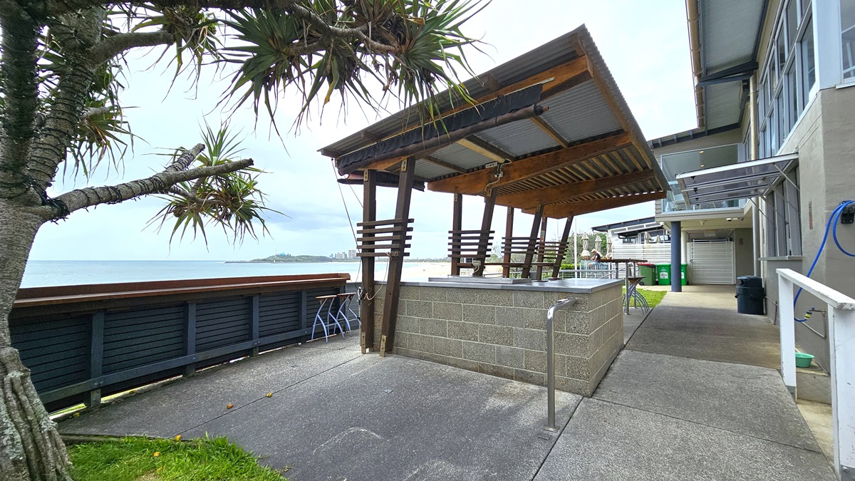 BBQ Breakfast Location - ANZAC Dawn Service - The Surf Club Mooloolaba. A coastal scene with a modern outdoor barbecue area in the centre, featuring a wooden and metal roof structure. To the left, a tree with spiky leaves is near a wooden railing overlooking the sea. In the background, distant land is visible across the water. On the right, a building with large windows and a concrete path leading to it. The ground is paved with concrete, and there is a patch of grass near the tree.