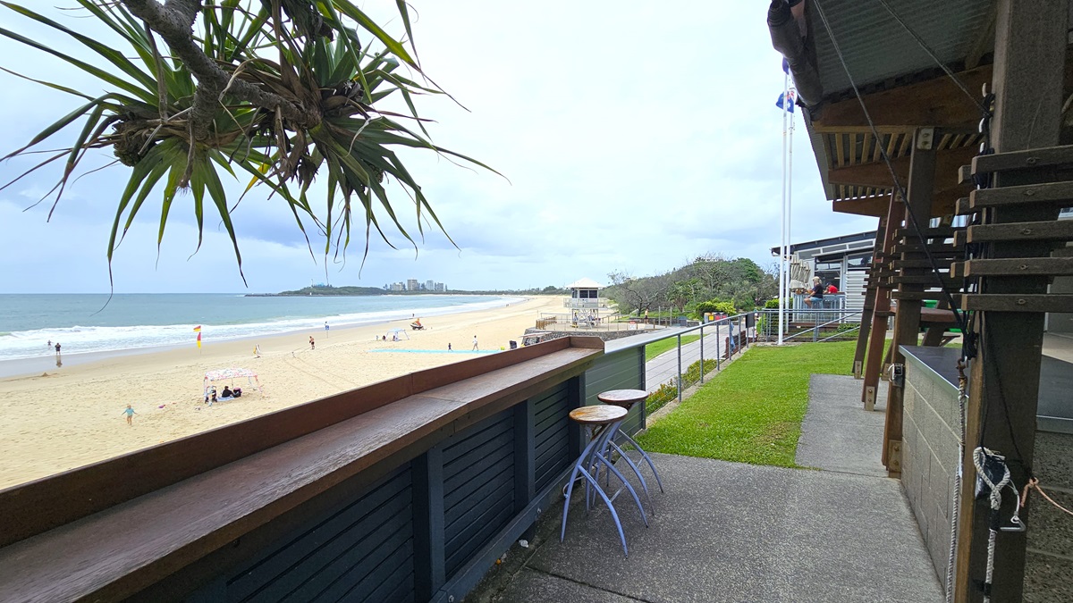 BBQ Breakfast Outlook - ANZAC Dawn Service - The Surf Club Mooloolaba. A coastal scene with a sandy beach stretching along the left side, where people are scattered enjoying the shore. The ocean waves gently lap against the sand. In the foreground, a wooden railing with a couple of bar stools overlooks the beach. On the right, a building with a sloped roof and a grassy area is visible. A tree with long, spiky leaves hangs overhead, partially framing the view.
