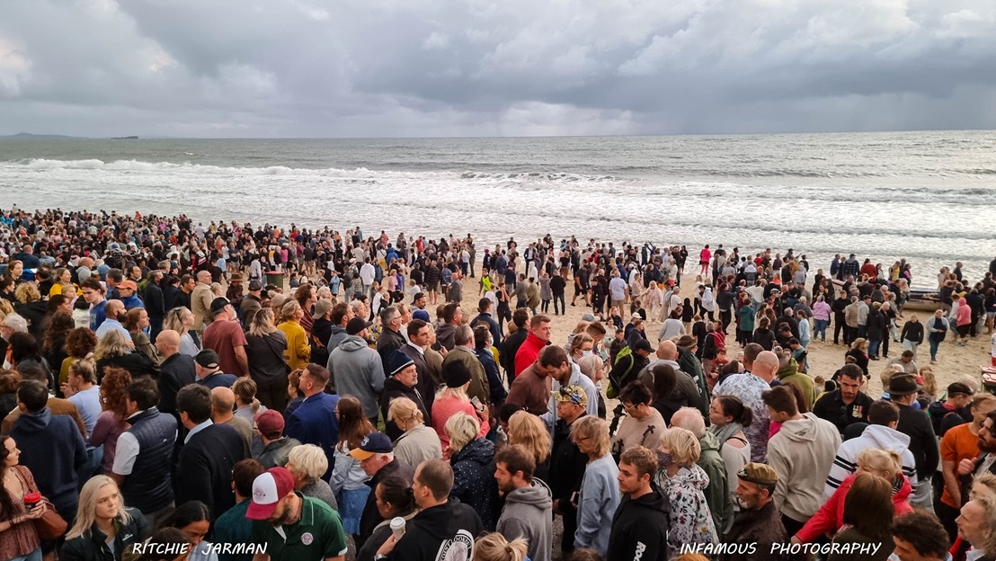 Attendees After Dawn - ANZAC Dawn Service - The Surf Club Mooloolaba. A crowded beach scene with a large number of people gathered on the sand. The ocean is in the background, with waves crashing onto the shore. The sky is overcast with grey clouds. People are dressed in casual clothing, and the crowd extends from the foreground to the water's edge.