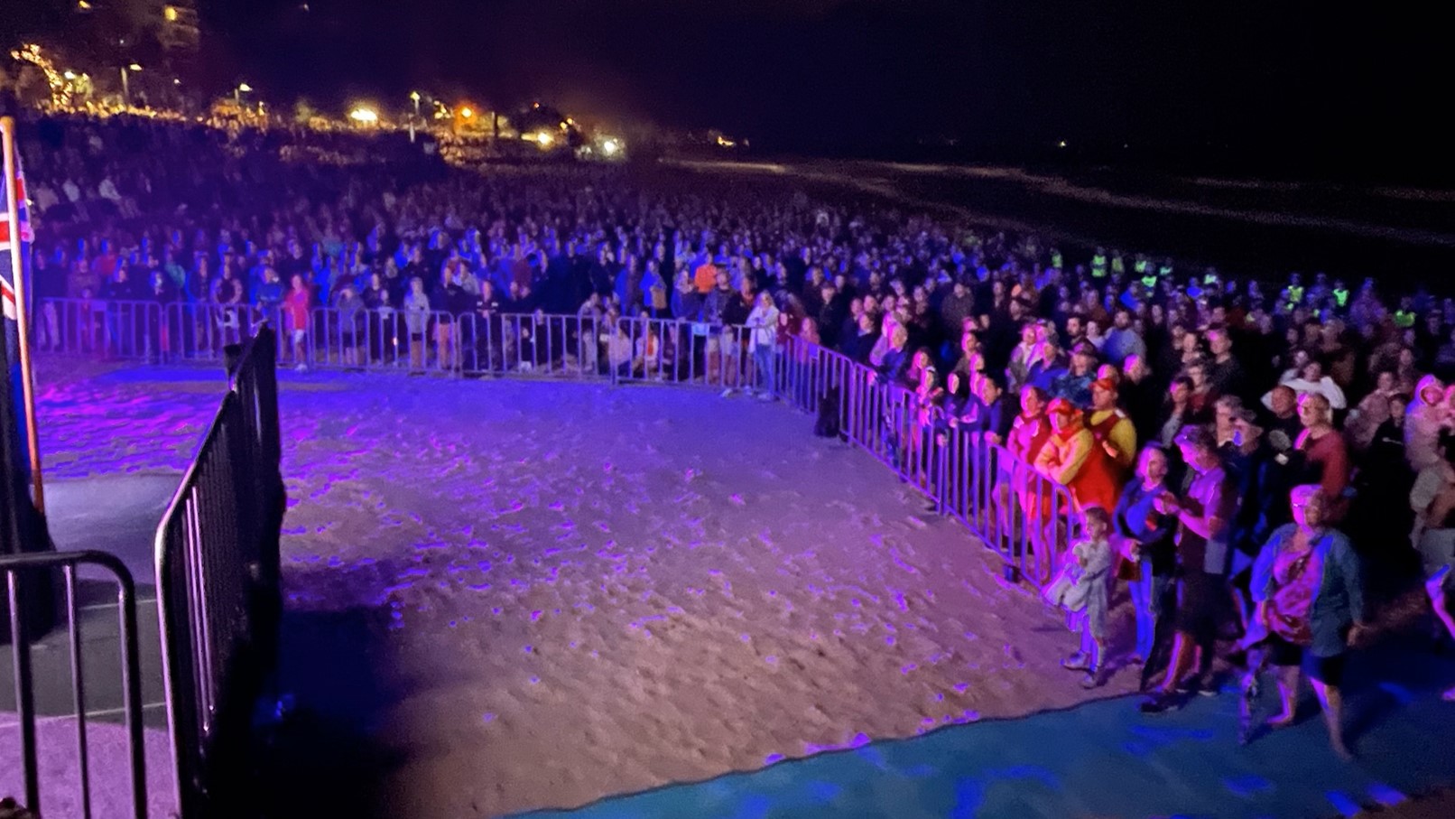 Attendees Before Dawn - ANZAC Dawn Service - The Surf Club Mooloolaba. A large crowd gathered on a sandy beach at night, illuminated by purple and blue lights. Metal barriers separate the audience from a stage area on the left, where part of a flag is visible. In the background, distant lights from buildings or street lamps are visible, creating a glowing effect. The people are dressed in various clothing, some in bright colours, and are standing closely together, watching the event.