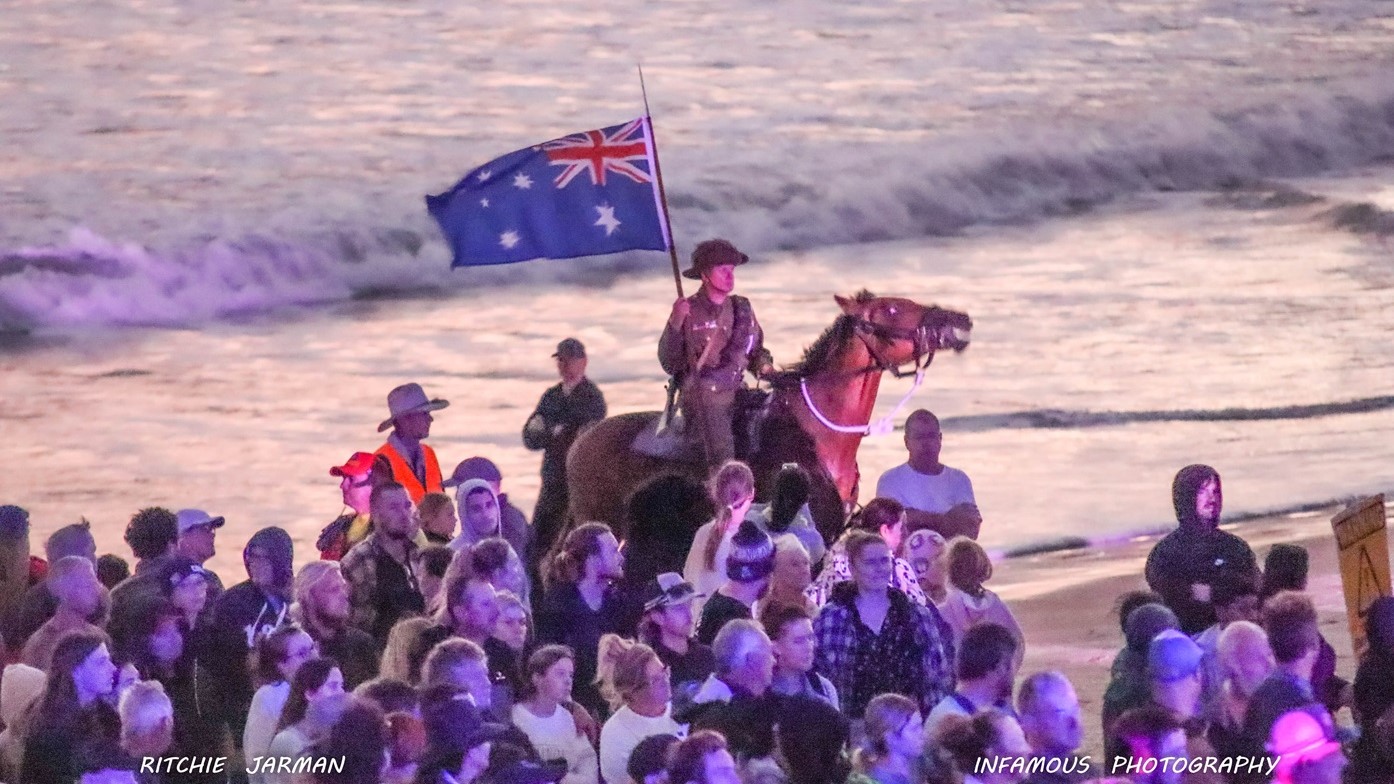 Attendees and Horse - ANZAC Dawn Service - The Surf Club Mooloolaba. A person on horseback is holding an Australian flag, riding along a beach. The horse is brown with a white bridle. In the foreground, a large crowd of people is gathered, some wearing hats and jackets. The ocean waves are visible in the background, with a soft, purple hue reflecting on the water. The scene is illuminated with a mix of natural and artificial light, creating a vibrant atmosphere.