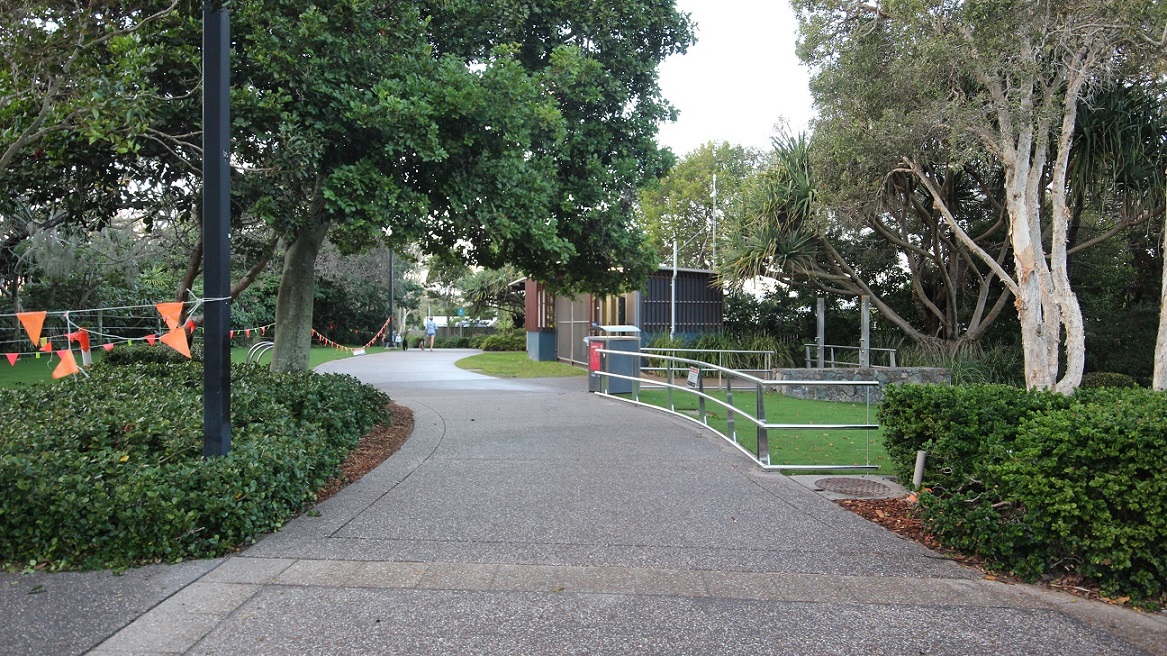 A curvy concrete pathway with a metal railing bordering the road on the right. Trimmed grass lines the roadside. To the left, a string of orange flags lines the road alongside trees and shrubs. In the distance, a low-rise structure with corrugated walls is visible. A curvy concrete pathway with a metal railing bordering the road on the right. Trimmed grass lines the roadside. To the left, a string of orange flags lines the road alongside trees and shrubs. In the distance, a low-rise structure with corrugated walls is visible.