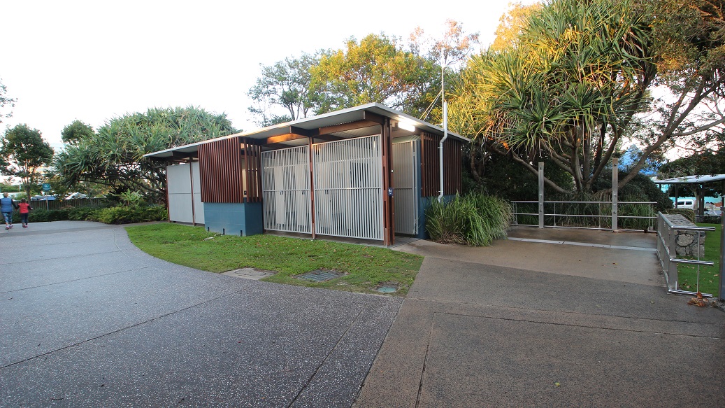 A low-rise structure with corrugated walls and metal railings. A metal fence, green grass, and trees border the road on the right. To the left is a curvy road with visible pedestrians. A low-rise structure with corrugated walls and metal railings. A metal fence, green grass, and trees border the road on the right. To the left is a curvy road with visible pedestrians.