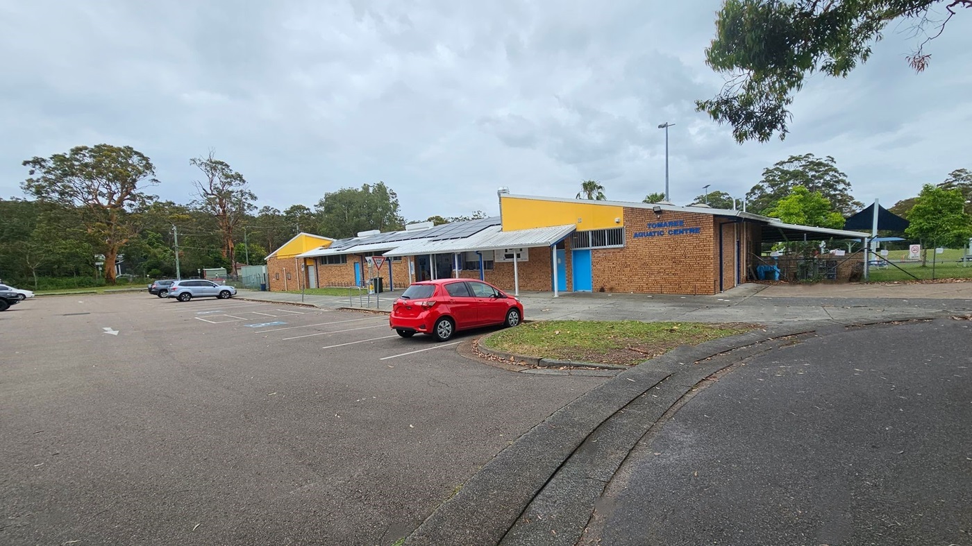 A brick building with a yellow and blue facade is on the right, labelled "Aquatic Centre". A red car is parked in front of it. To the left, there are several parking spaces with a few cars. The background features tall trees and greenery. The foreground shows a paved area with some scattered leaves. A brick building with a yellow and blue facade is on the right, labelled "Aquatic Centre". A red car is parked in front of it. To the left, there are several parking spaces with a few cars. The background features tall trees and greenery. The foreground shows a paved area with some scattered leaves.