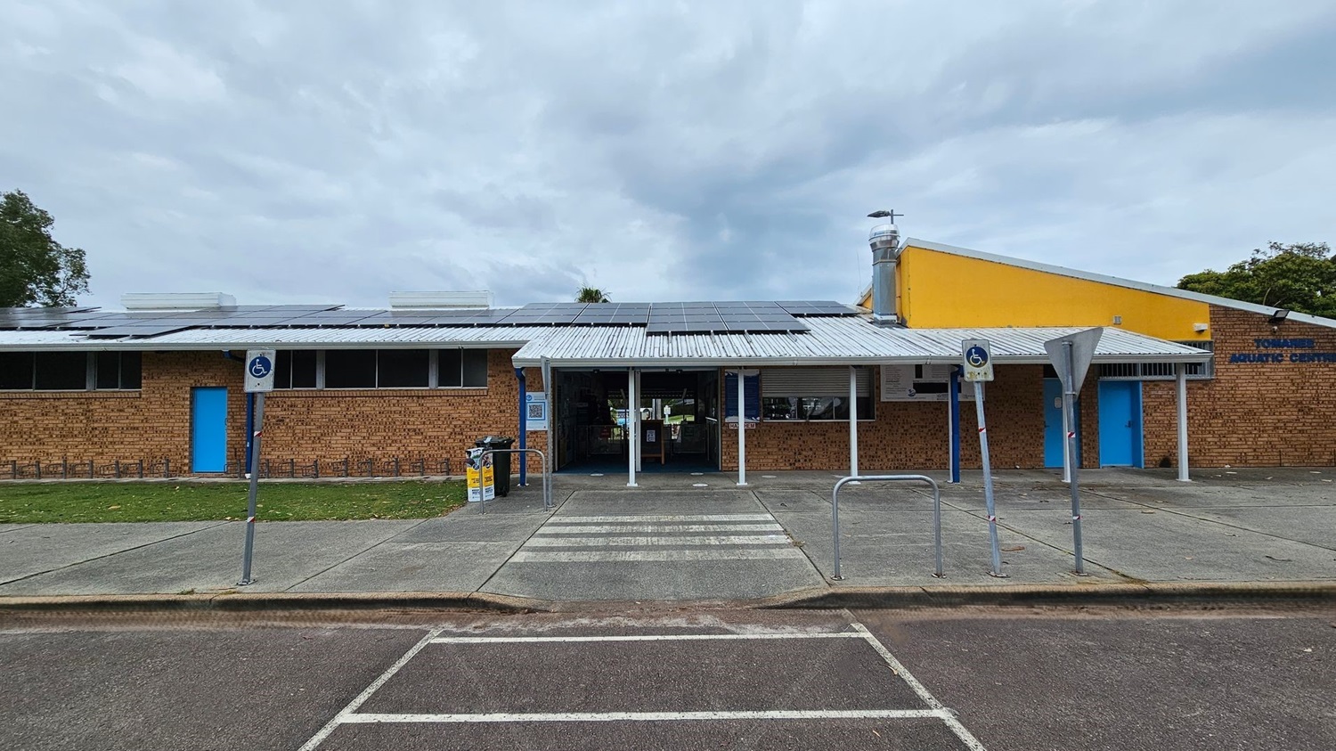 A brick building with a metal roof featuring solar panels. The entrance is in the centre, flanked by blue doors on either side. A yellow section of the building is on the right. There are signs for disabled parking in front, with a zebra crossing leading to the entrance. The foreground has a paved area and parking spaces, with some fallen leaves scattered around. Trees are visible in the background. A brick building with a metal roof featuring solar panels. The entrance is in the centre, flanked by blue doors on either side. A yellow section of the building is on the right. There are signs for disabled parking in front, with a zebra crossing leading to the entrance. The foreground has a paved area and parking spaces, with some fallen leaves scattered around. Trees are visible in the background.