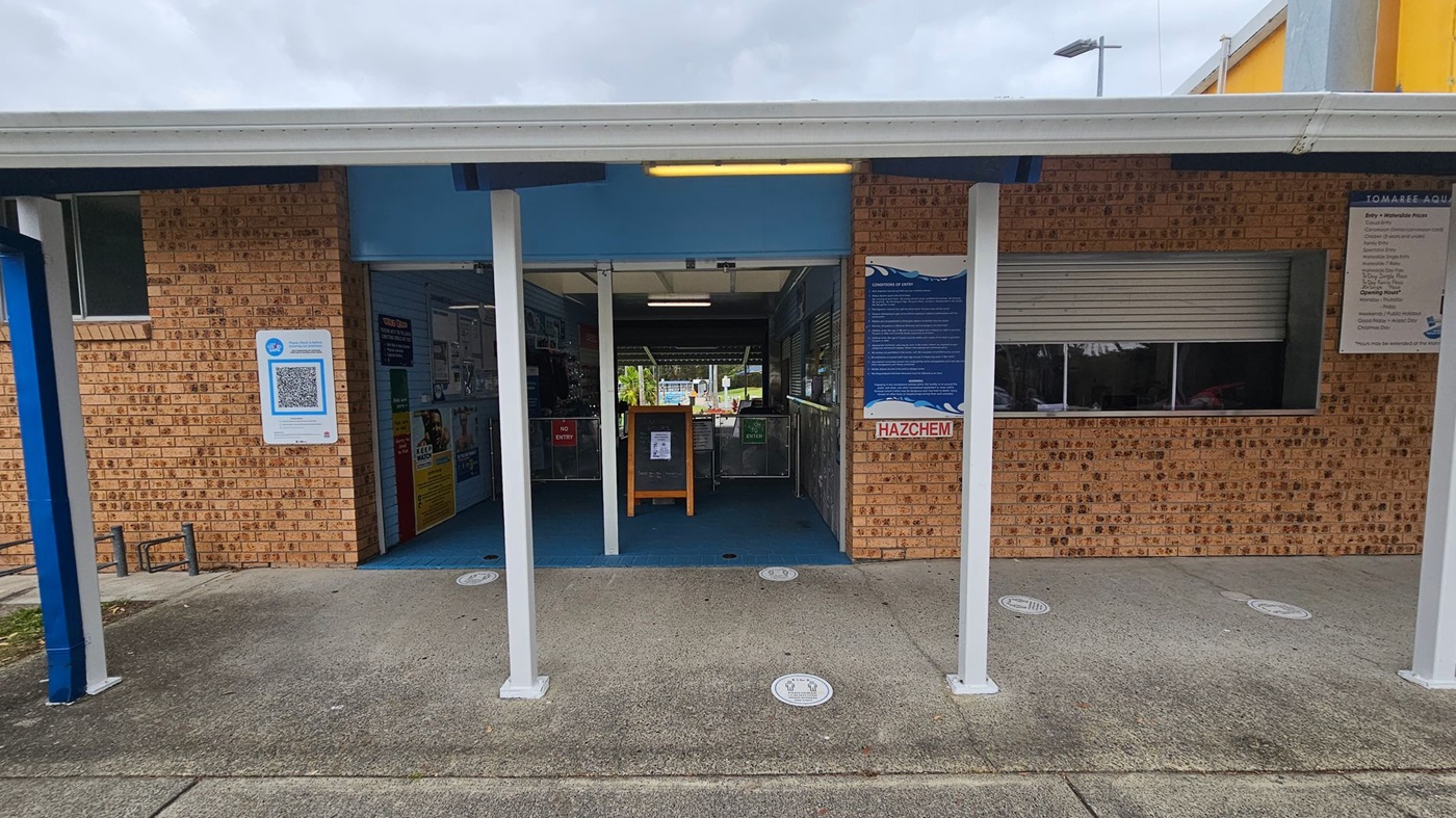 A brick building with a covered entrance supported by white pillars. On the left, a QR code sign is displayed on the wall. The entrance is open, revealing a blue interior with various signs and a wooden stand. To the right, a "HAZCHEM" sign and a shuttered window are visible. The ground is a mix of green and grey concrete, and there are circular markers on the floor. A sign with information is mounted on the right wall. A brick building with a covered entrance supported by white pillars. On the left, a QR code sign is displayed on the wall. The entrance is open, revealing a blue interior with various signs and a wooden stand. To the right, a "HAZCHEM" sign and a shuttered window are visible. The ground is a mix of green and grey concrete, and there are circular markers on the floor. A sign with information is mounted on the right wall.