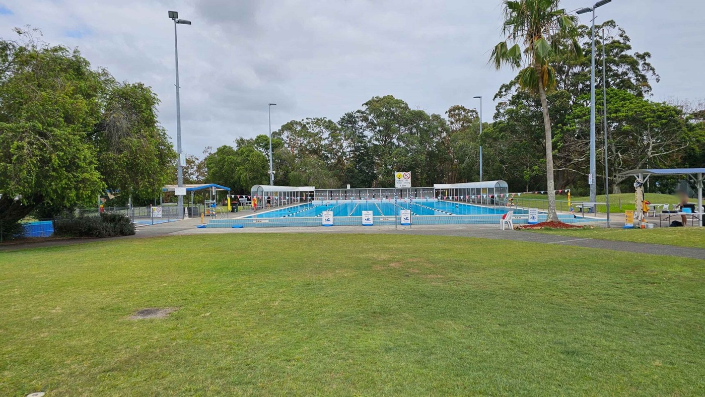 A park with a concrete path leading towards a swimming pool area. On the left, there is a large blue water slide structure surrounded by trees. In the centre, two tall palm trees stand near the path. On the right, a pool is covered by dark triangular shade sails, with signs and benches nearby. The grass is green and well-maintained, and there are landscaped flower beds near the path. Trees line the background. A park with a concrete path leading towards a swimming pool area. On the left, there is a large blue water slide structure surrounded by trees. In the centre, two tall palm trees stand near the path. On the right, a pool is covered by dark triangular shade sails, with signs and benches nearby. The grass is green and well-maintained, and there are landscaped flower beds near the path. Trees line the background.