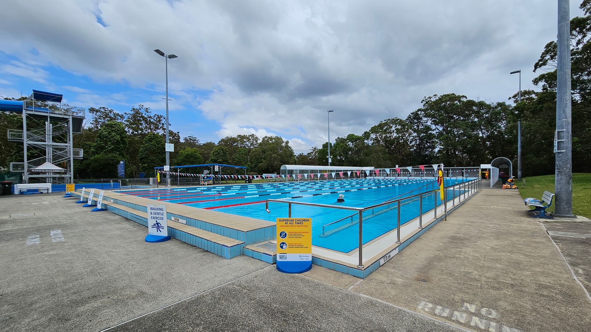 An outdoor swimming pool with marked lanes is in the centre, surrounded by a tiled deck. On the left, there's a tall water slide structure with stairs. Signs around the pool indicate safety rules. On the right, a pathway with a "No Running" warning leads to a covered area. Benches and tall lights are visible, with trees in the background. The ground is a mix of concrete and grass. An outdoor swimming pool with marked lanes is in the centre, surrounded by a tiled deck. On the left, there's a tall water slide structure with stairs. Signs around the pool indicate safety rules. On the right, a pathway with a "No Running" warning leads to a covered area. Benches and tall lights are visible, with trees in the background. The ground is a mix of concrete and grass.