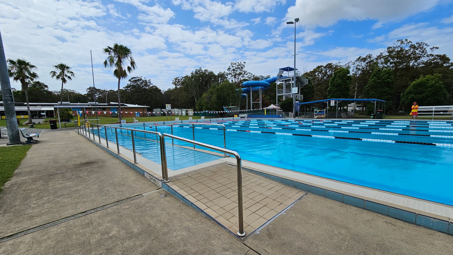 Outdoor swimming pool with marked lanes on the right, surrounded by a metal railing. A lifeguard in a red and yellow uniform stands near the pool. On the left, a paved walkway with benches and a grassy area. In the background, there are palm trees, a building with a low roof, and a blue water slide structure. Tall trees are visible beyond the pool area. Outdoor swimming pool with marked lanes on the right, surrounded by a metal railing. A lifeguard in a red and yellow uniform stands near the pool. On the left, a paved walkway with benches and a grassy area. In the background, there are palm trees, a building with a low roof, and a blue water slide structure. Tall trees are visible beyond the pool area.