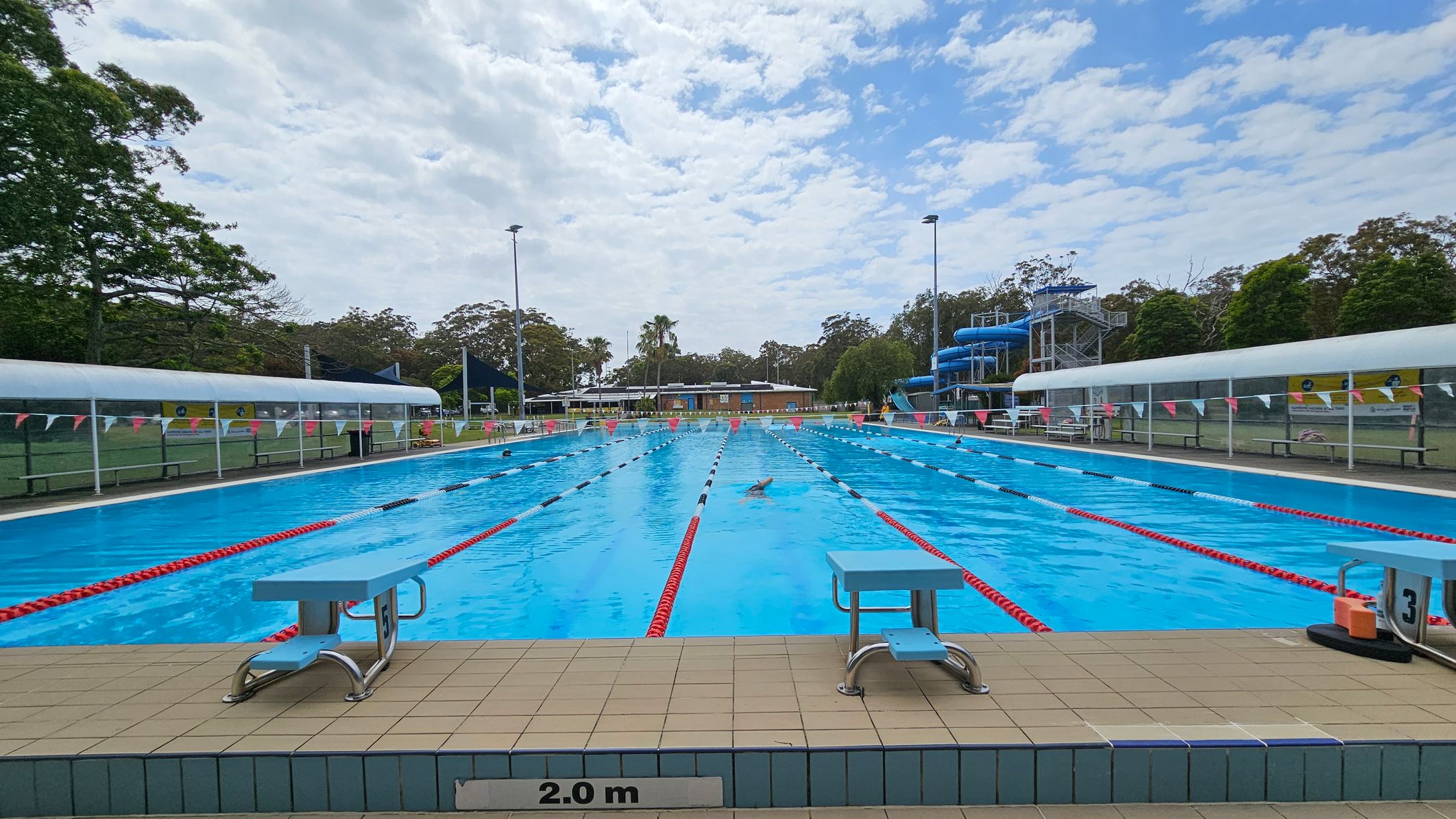 An outdoor swimming pool with clear blue water, marked with red and white lane dividers. Two diving boards are positioned at the pool's edge, with a depth marker indicating 2.0 metres. On the left and right, there are covered seating areas with benches. In the background, a blue water slide structure is visible on the right, surrounded by trees and a grassy area. A few swimmers are in the pool, and a building is seen in the distance. An outdoor swimming pool with clear blue water, marked with red and white lane dividers. Two diving boards are positioned at the pool's edge, with a depth marker indicating 2.0 metres. On the left and right, there are covered seating areas with benches. In the background, a blue water slide structure is visible on the right, surrounded by trees and a grassy area. A few swimmers are in the pool, and a building is seen in the distance.