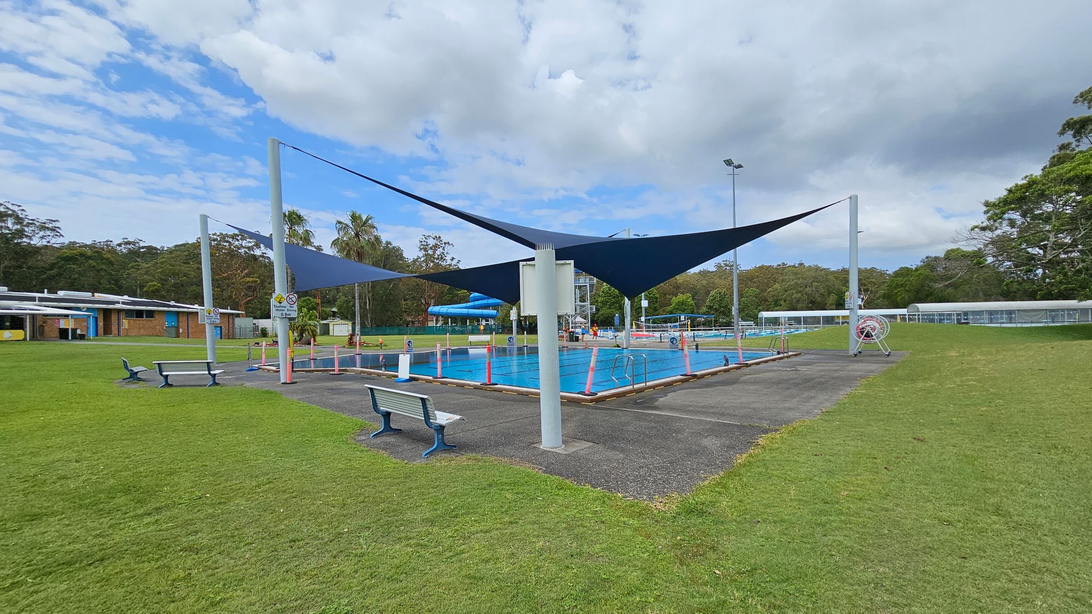 A swimming pool is in the centre, surrounded by a paved area with benches. Blue shade sails are suspended above the pool. On the left, there is a building with a blue and yellow facade. The right side features grassy areas and a covered walkway. Trees and greenery are visible in the background. A swimming pool is in the centre, surrounded by a paved area with benches. Blue shade sails are suspended above the pool. On the left, there is a building with a blue and yellow facade. The right side features grassy areas and a covered walkway. Trees and greenery are visible in the background.