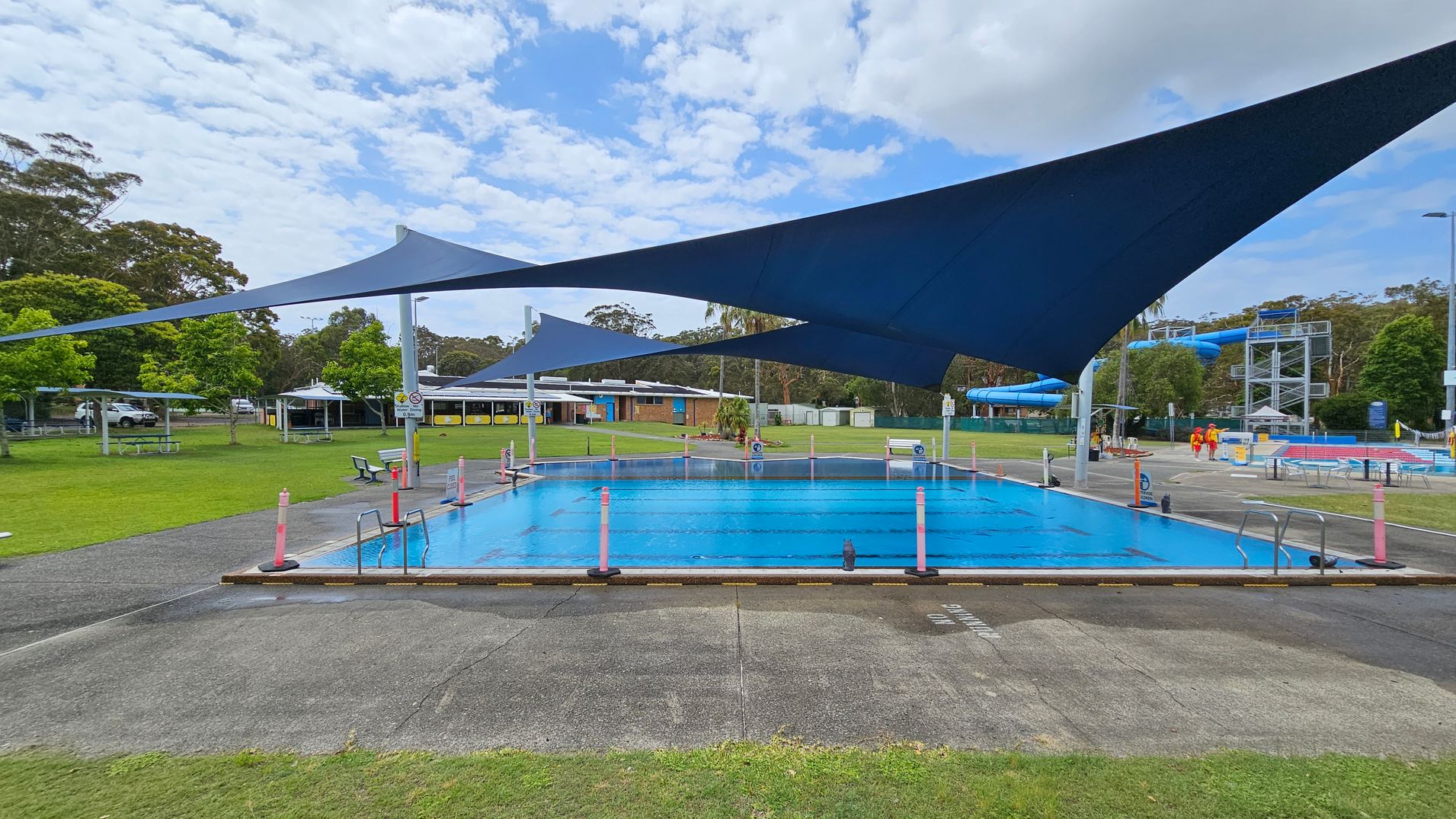 A swimming pool with blue water is in the centre, surrounded by a concrete path. Above, large blue shade sails provide cover. On the left, there are green lawns with picnic tables and trees. In the background, a building with a white roof is visible. On the right, two people in red and yellow uniforms walk near a blue water slide structure. The area is open and surrounded by greenery. A swimming pool with blue water is in the centre, surrounded by a concrete path. Above, large blue shade sails provide cover. On the left, there are green lawns with picnic tables and trees. In the background, a building with a white roof is visible. On the right, two people in red and yellow uniforms walk near a blue water slide structure. The area is open and surrounded by greenery.