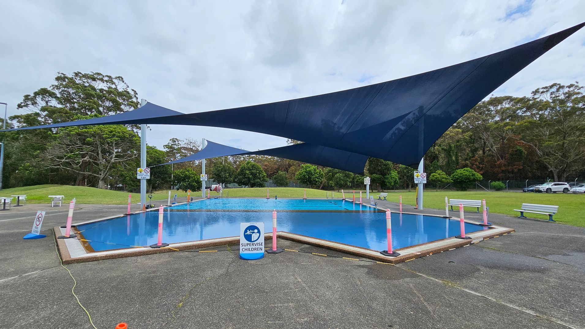 A small outdoor pool is surrounded by a paved area with a sign reading "Supervise Children" at the front. The pool is covered by large, dark blue shade sails. Pink bollards are placed around the pool's perimeter. Benches are positioned on the grass in the background, with trees and a parked car visible beyond. The ground is a mix of concrete and grass. A small outdoor pool is surrounded by a paved area with a sign reading "Supervise Children" at the front. The pool is covered by large, dark blue shade sails. Pink bollards are placed around the pool's perimeter. Benches are positioned on the grass in the background, with trees and a parked car visible beyond. The ground is a mix of concrete and grass.