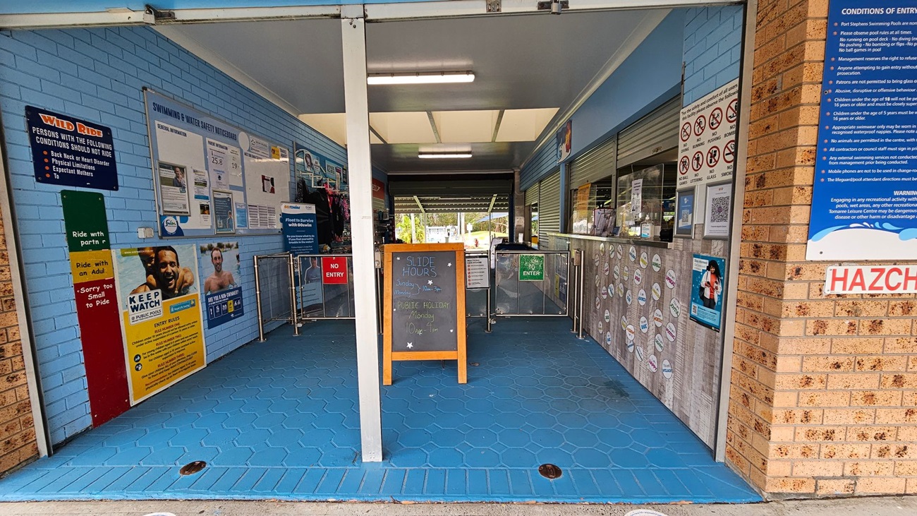 Entrance to a swimming facility with blue hexagonal floor tiles. On the left, a blue wall displays various safety and informational signs, including a "Wild Ride" notice. In the centre, a wooden board lists slide hours. On the right, a brick wall with a "Conditions of Entry" sign and a counter with closed shutters. Above, fluorescent lights illuminate the area. The entrance is flanked by metal barriers with "No Entry" and "Enter" signs. Entrance to a swimming facility with blue hexagonal floor tiles. On the left, a blue wall displays various safety and informational signs, including a "Wild Ride" notice. In the centre, a wooden board lists slide hours. On the right, a brick wall with a "Conditions of Entry" sign and a counter with closed shutters. Above, fluorescent lights illuminate the area. The entrance is flanked by metal barriers with "No Entry" and "Enter" signs.