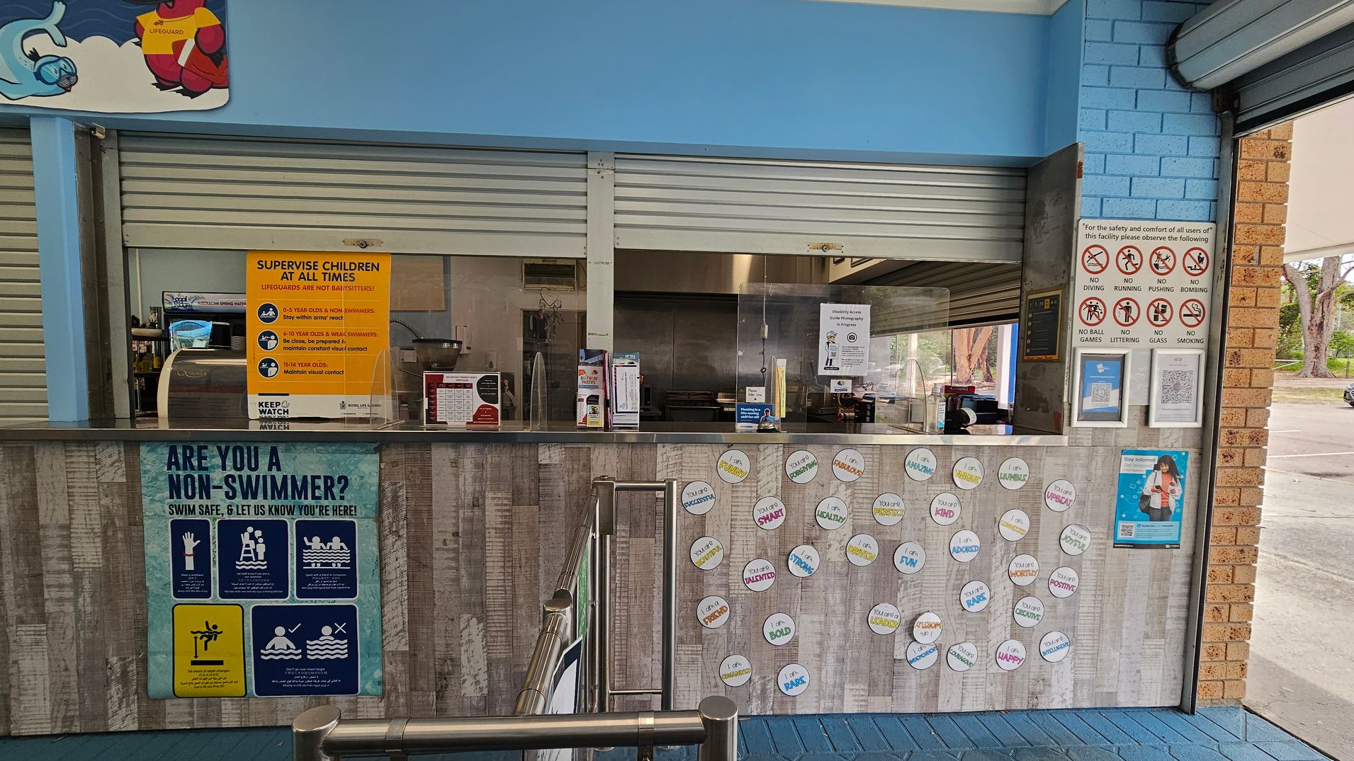A counter area with a blue and brick wall background. The counter has various signs, including a large yellow one about supervising children and a blue one asking if you are a non-swimmer. Circular stickers with positive affirmations are displayed on the front. Behind the counter, there are closed metal shutters and a small kitchen area. On the right, there are safety signs prohibiting activities like diving and running. The floor is tiled in blue. A counter area with a blue and brick wall background. The counter has various signs, including a large yellow one about supervising children and a blue one asking if you are a non-swimmer. Circular stickers with positive affirmations are displayed on the front. Behind the counter, there are closed metal shutters and a small kitchen area. On the right, there are safety signs prohibiting activities like diving and running. The floor is tiled in blue.