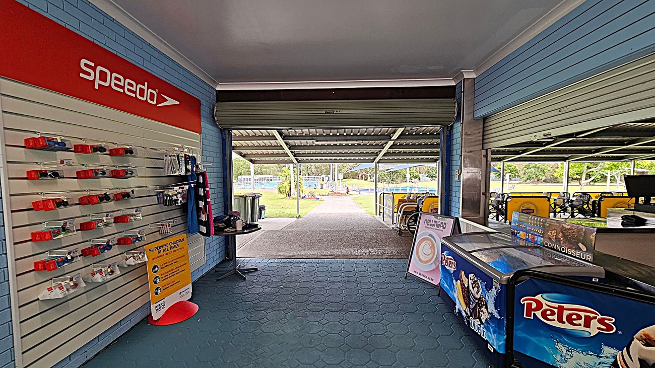 On the left, a display wall with Speedo-branded swimming accessories and a sign about supervising children. In the middle, an open area leads to an outdoor space with a path and greenery. On the right, a counter with a Peters ice cream freezer and a menu board. The floor is covered with hexagonal tiles, and the ceiling is plain. On the left, a display wall with Speedo-branded swimming accessories and a sign about supervising children. In the middle, an open area leads to an outdoor space with a path and greenery. On the right, a counter with a Peters ice cream freezer and a menu board. The floor is covered with hexagonal tiles, and the ceiling is plain.
