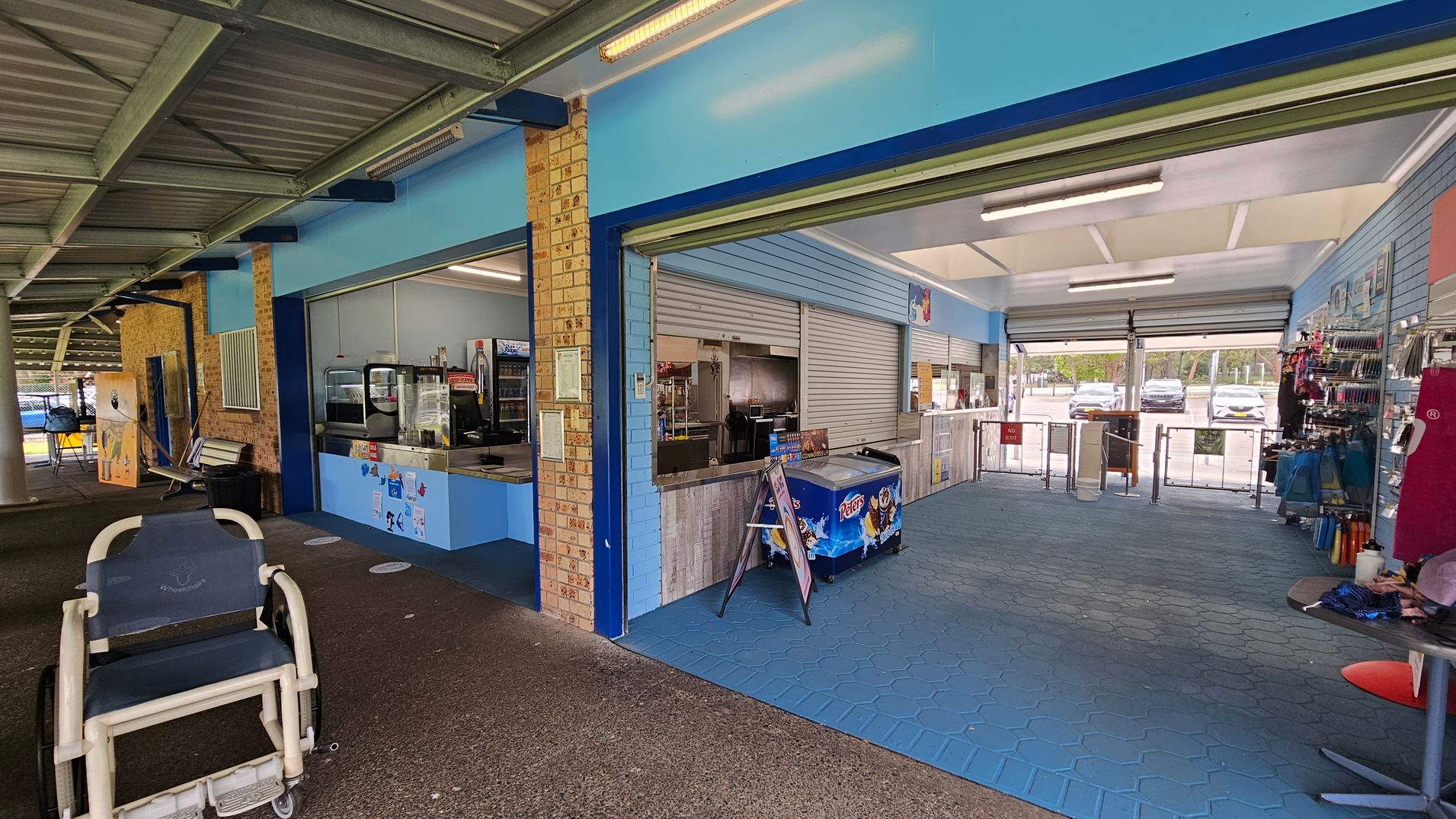 A covered outdoor area with a corrugated metal ceiling and a concrete floor. On the left, there's a wheelchair and a bench against a brick wall. In the centre, a kiosk with a blue counter displays a variety of items, including an ice cream freezer. The right side has a blue-tiled floor and shelves with merchandise. The background shows a car park visible through open roller doors. A covered outdoor area with a corrugated metal ceiling and a concrete floor. On the left, there's a wheelchair and a bench against a brick wall. In the centre, a kiosk with a blue counter displays a variety of items, including an ice cream freezer. The right side has a blue-tiled floor and shelves with merchandise. The background shows a car park visible through open roller doors.